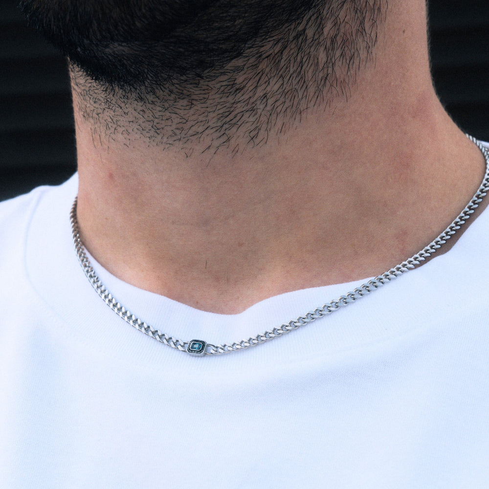 Close-up of a person wearing a silver chain necklace with a small green pendant on a white background.