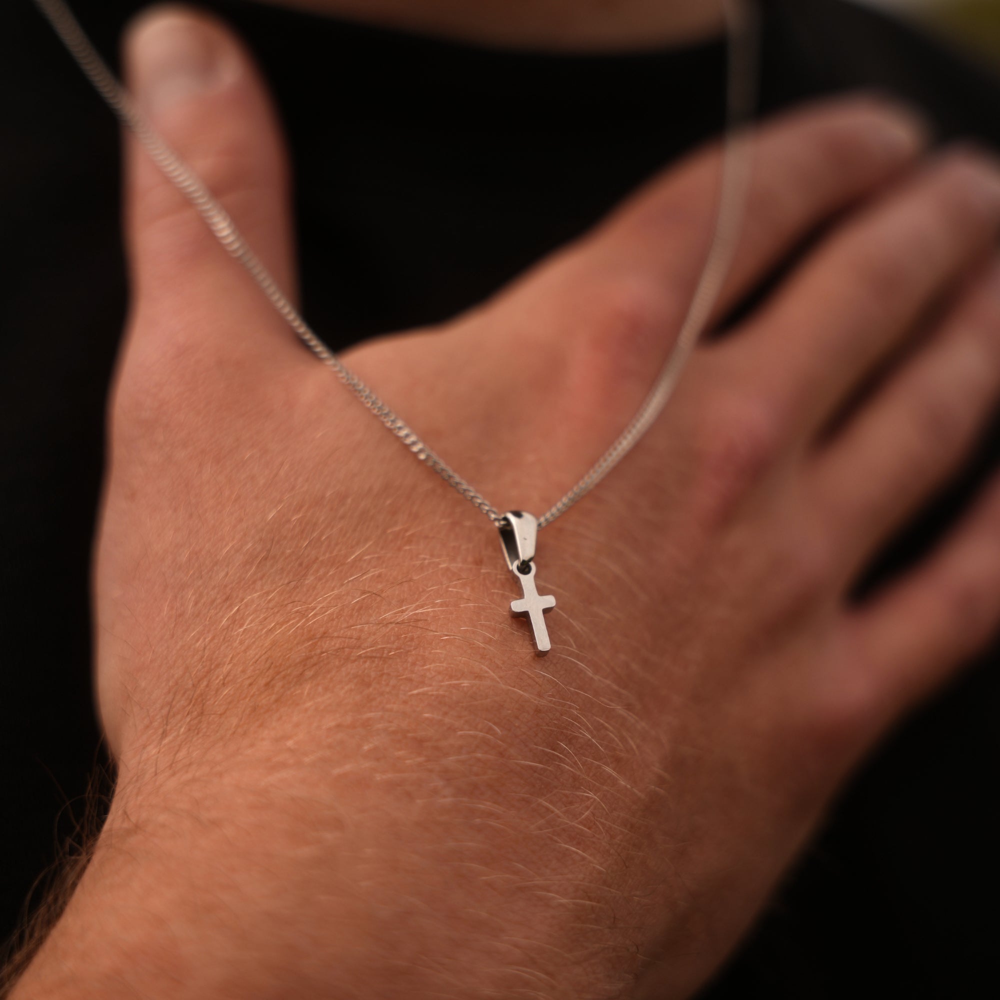 Hand wearing a silver necklace with a cross pendant on a dark background