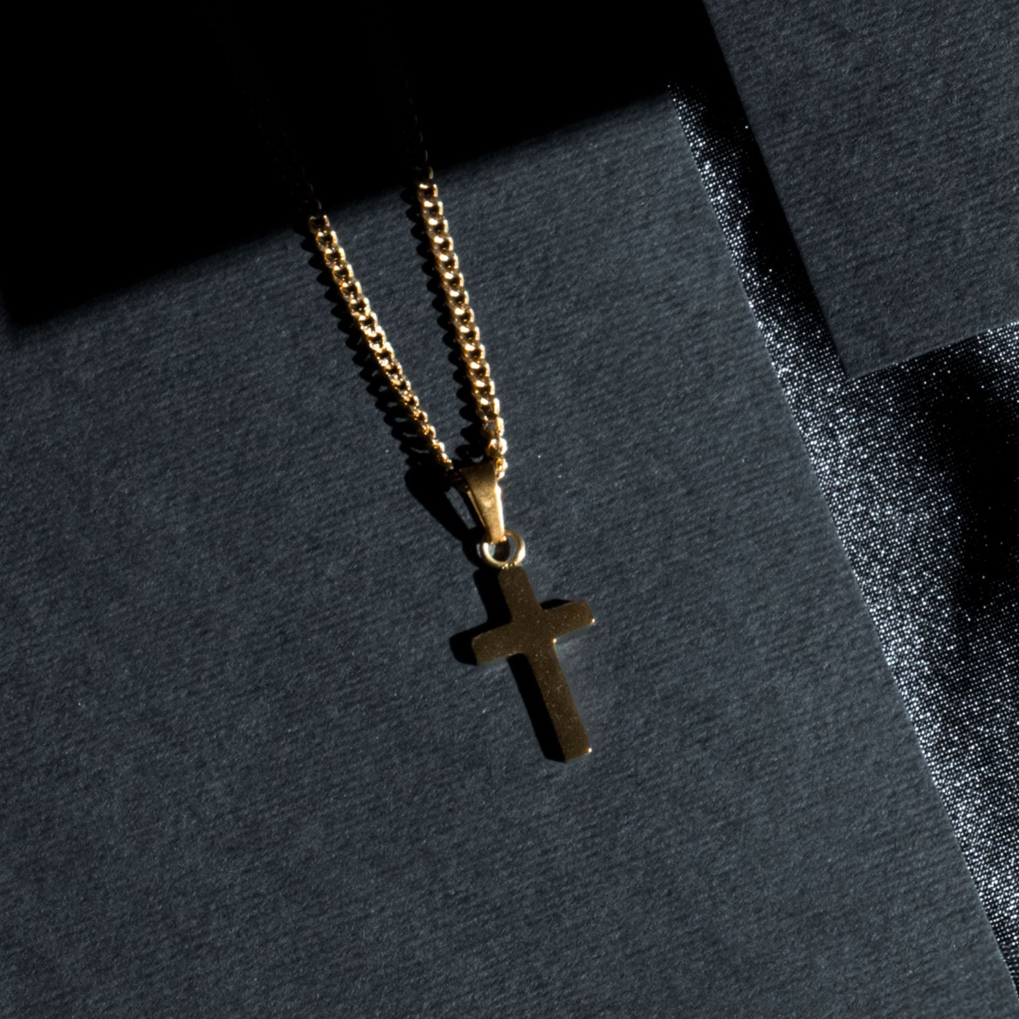 A gold cross pendant necklace on a gold chain, displayed on a textured dark gray surface with dramatic shadows.