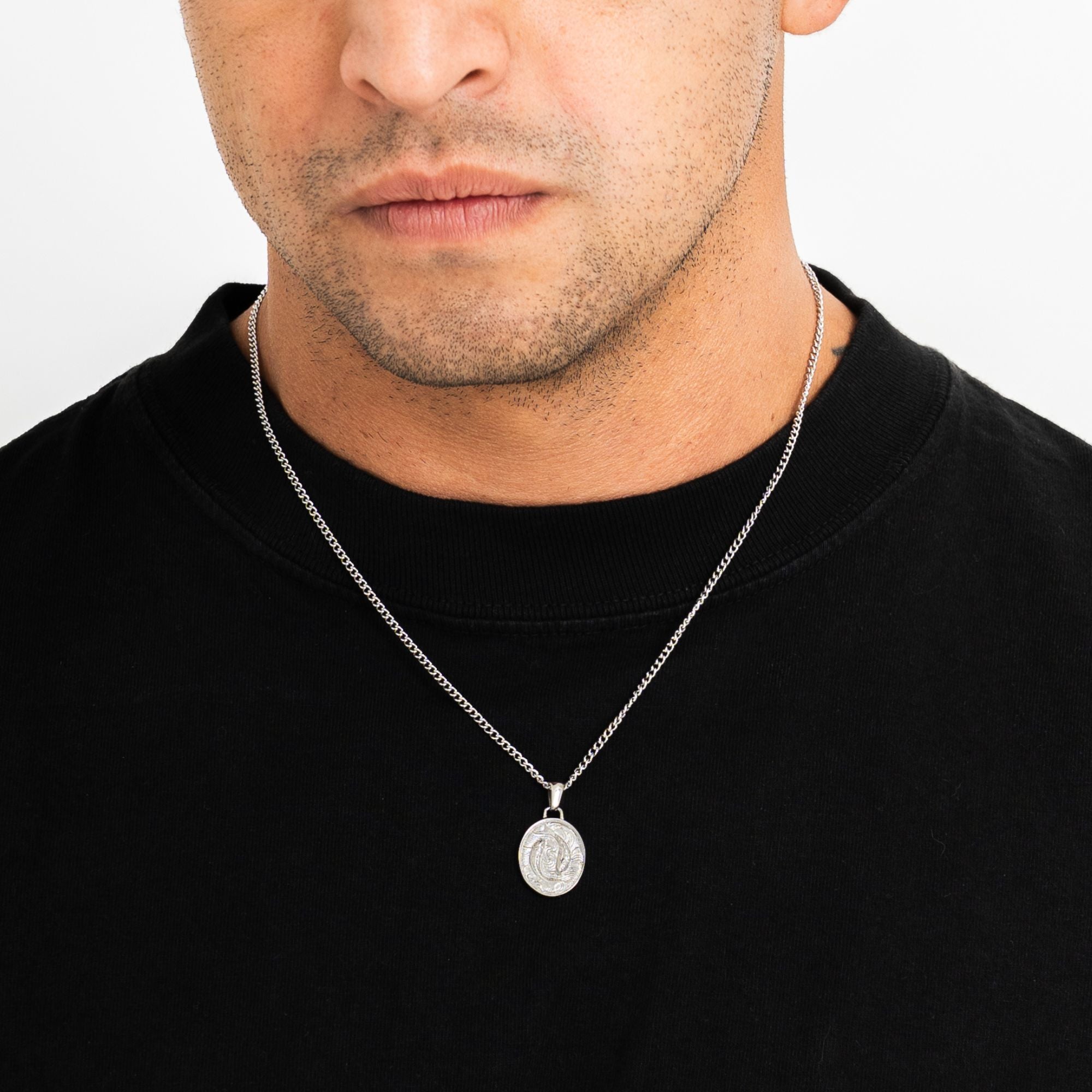 A close-up of a man wearing a silver chain necklace with a round medallion pendant, shown against a black crewneck shirt and a light gray background.