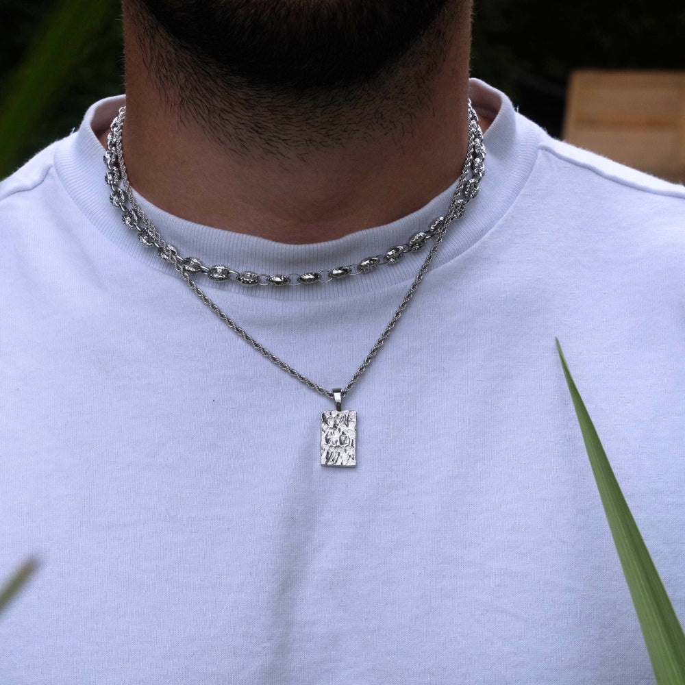 Person wearing a silver chain necklace with a rectangular pendant against a blurred natural background