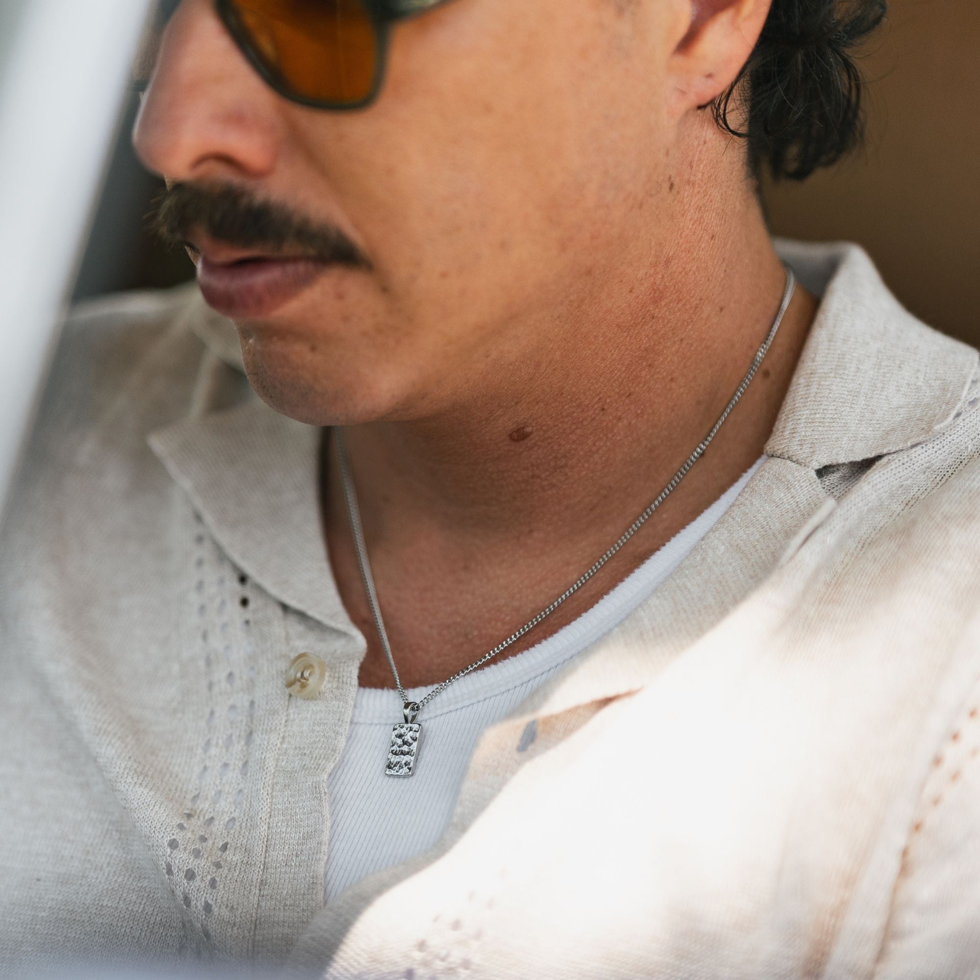 A close-up lifestyle shot of a man wearing a silver chain necklace with a small skull pendant, paired with a light beige linen polo shirt.