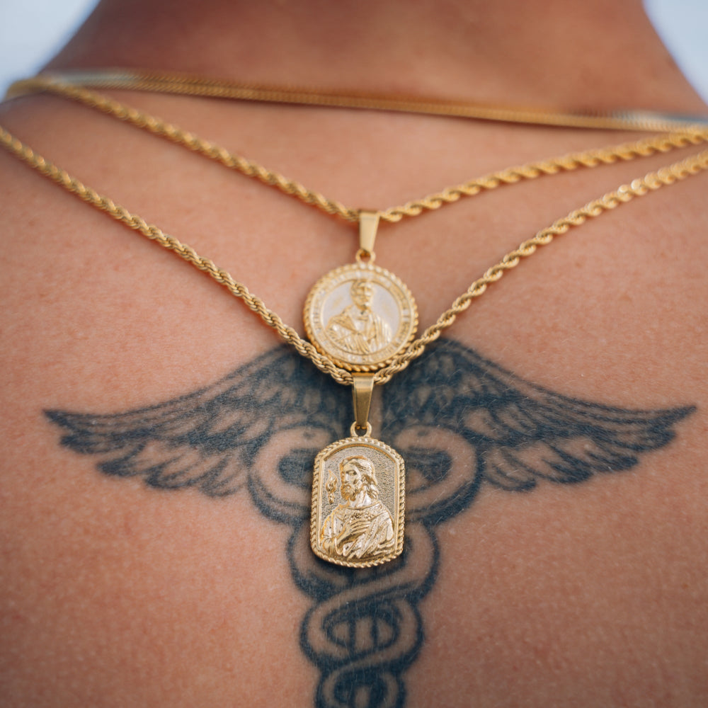 Gold necklaces with pendants on a person's neck with a tattoo of a caduceus.