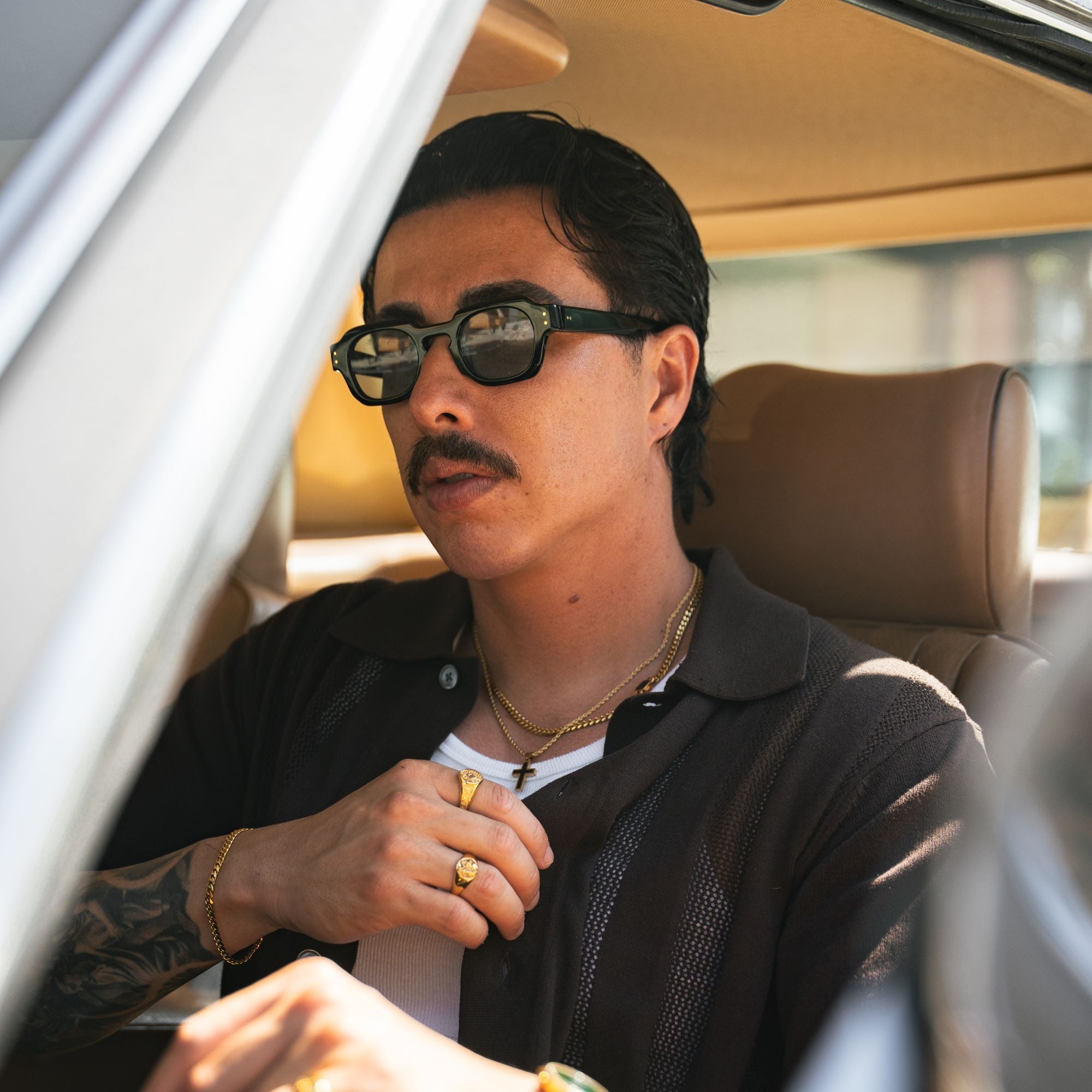 A man wearing black sunglasses, a black textured polo shirt, and gold jewelry sitting inside a car with beige leather seats.