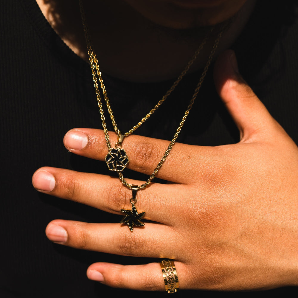 Hand wearing a gold ring with a gold chain featuring a hexagonal pendant against a black background