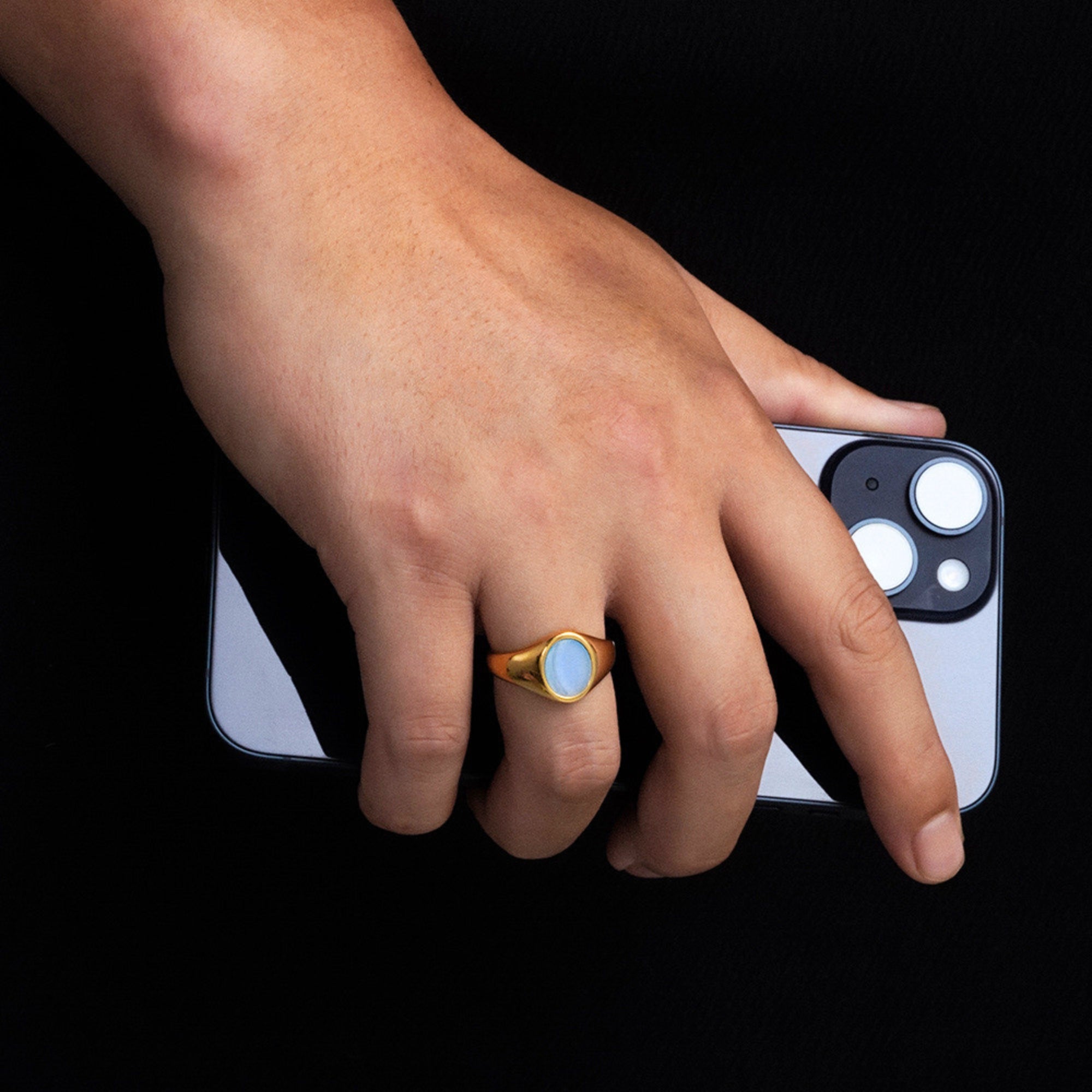 Hand holding a phone with a gold ring featuring a blue stone on a black background