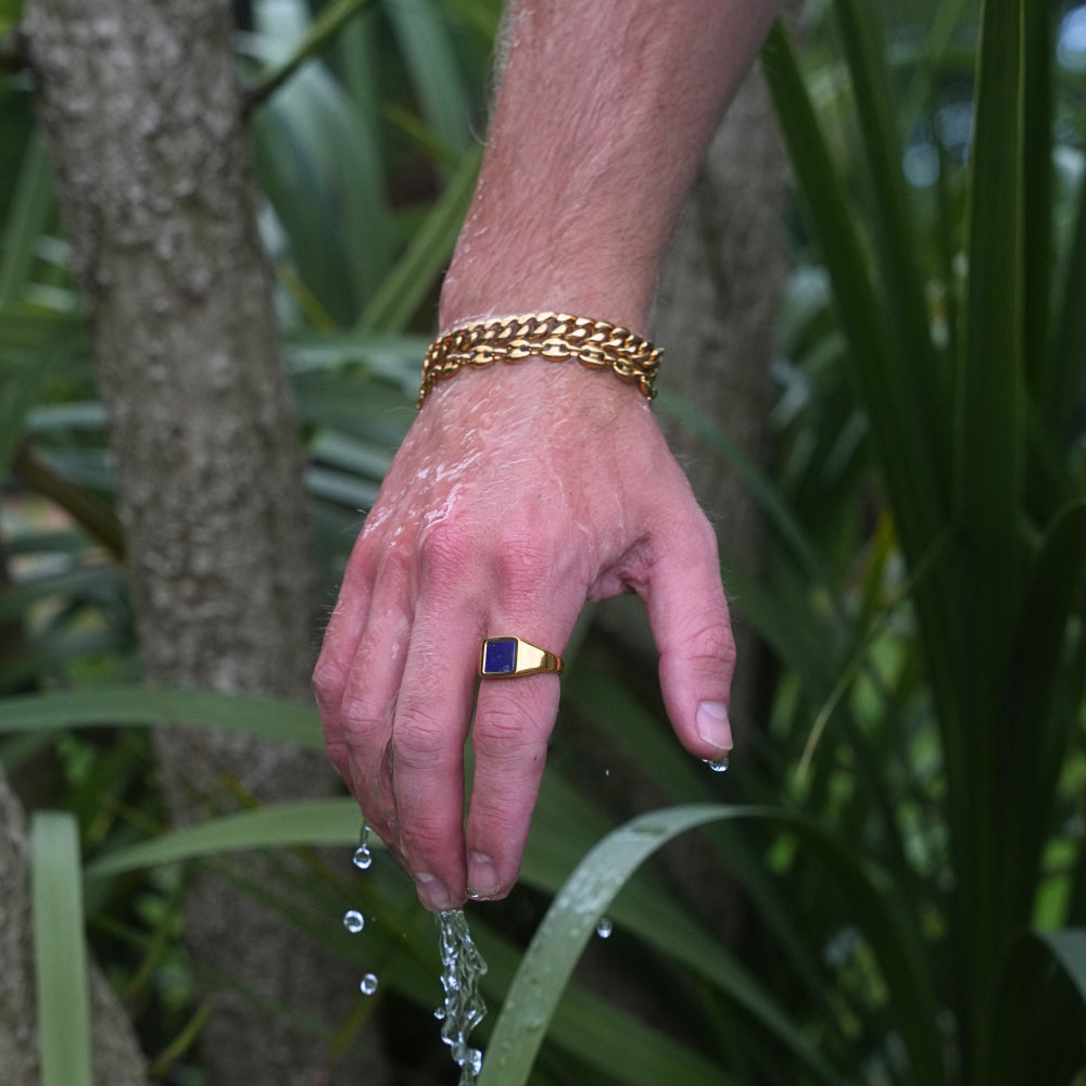 Hand with a gold ring and bracelet holding water droplets against a natural background