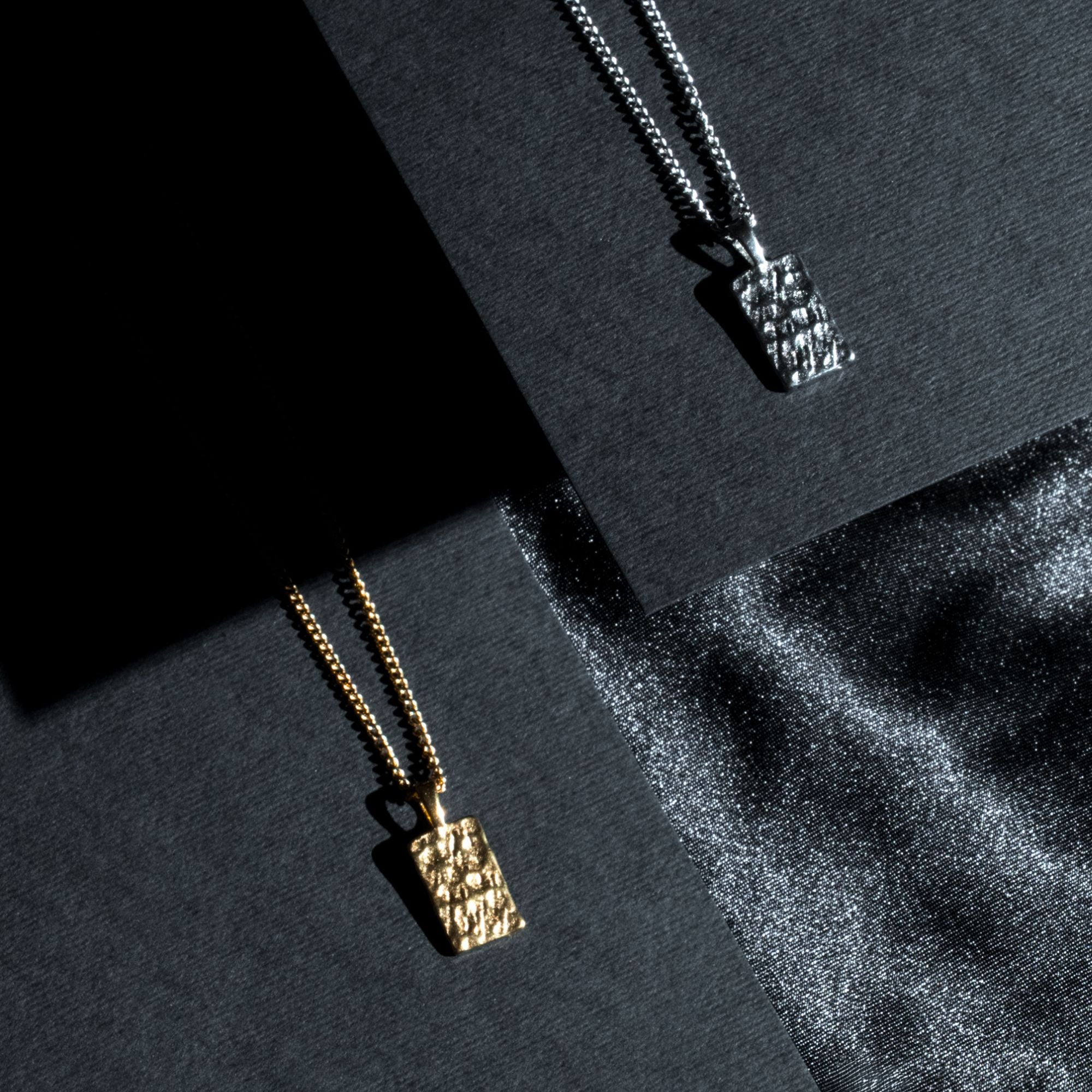 Two textured rectangular pendant necklaces on black display cards, one gold-toned and one silver-toned, with dramatic shadow lighting and a sparkly black fabric surface.