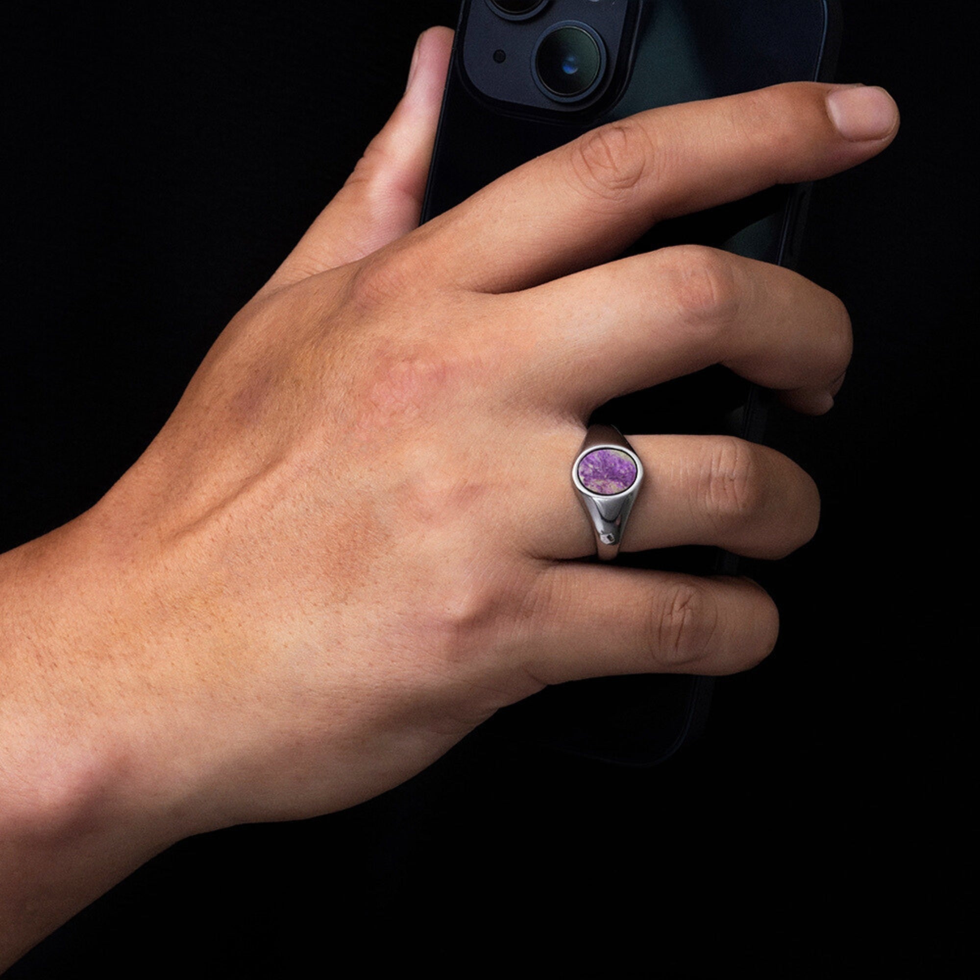 Hand wearing a silver ring with a purple gemstone holding a smartphone on a black background