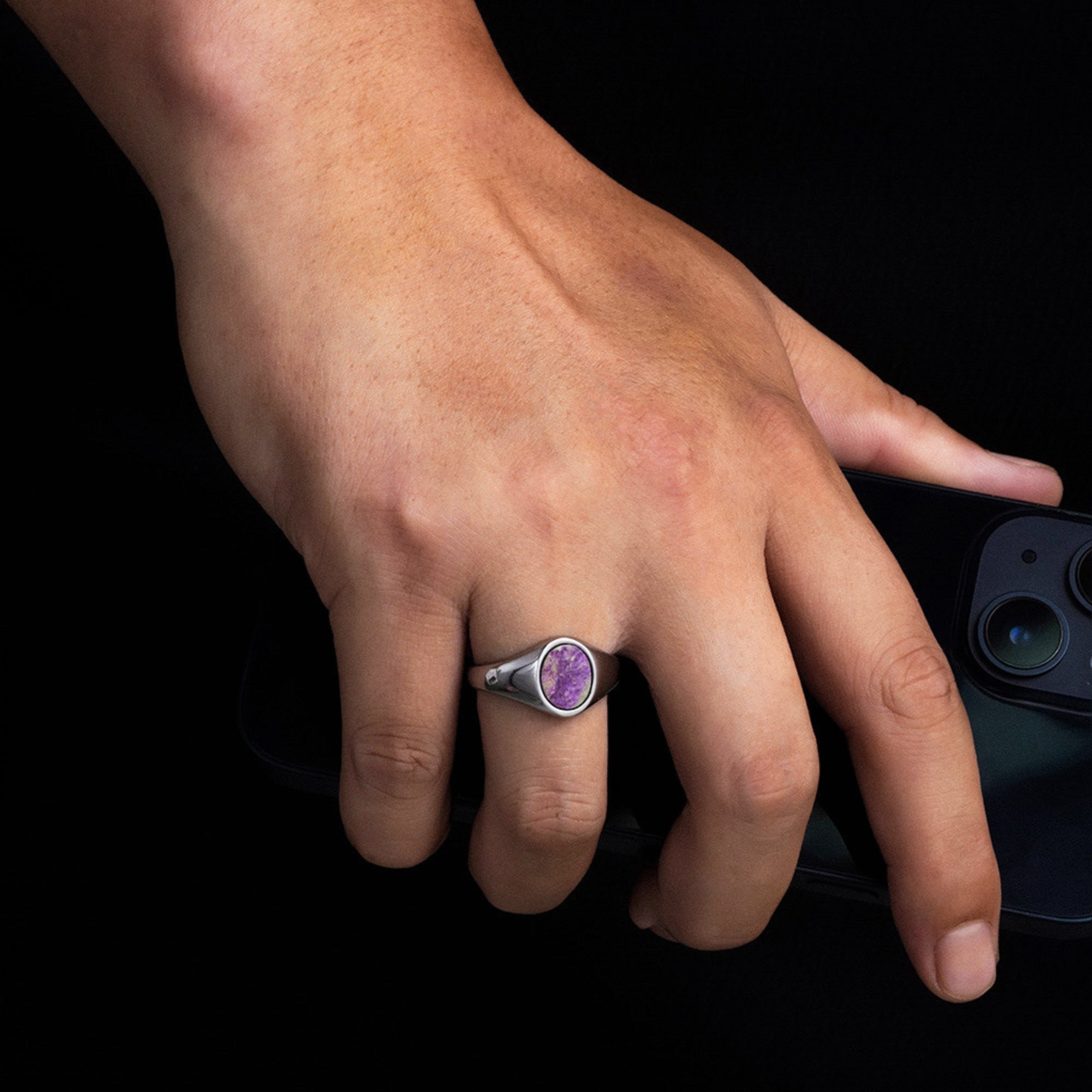Hand wearing a ring with a purple gemstone holding a smartphone on a black background