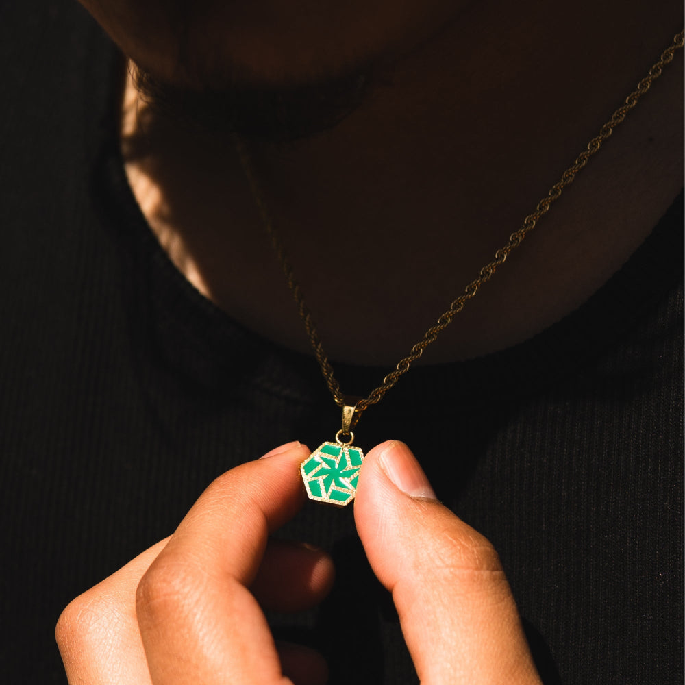 Person holding a gold necklace with a green hexagonal pendant against a dark background