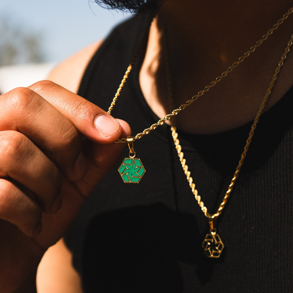 Person wearing two gold necklaces with green and gold pendants against a blurred outdoor background