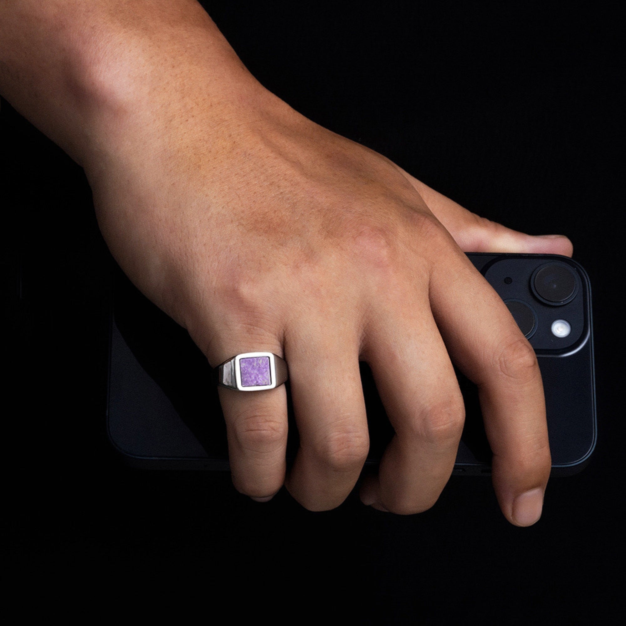 Hand holding a smartphone with a purple gemstone ring on a black background