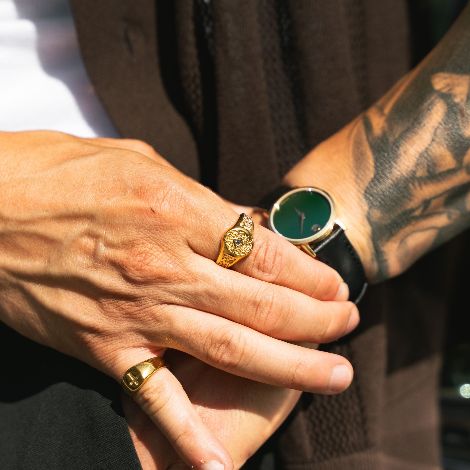 A close-up lifestyle shot of a person's hands and wrist, showcasing gold jewelry. The most prominent product is a gold signet ring with a detailed crest design on the top, worn on the ring finger.