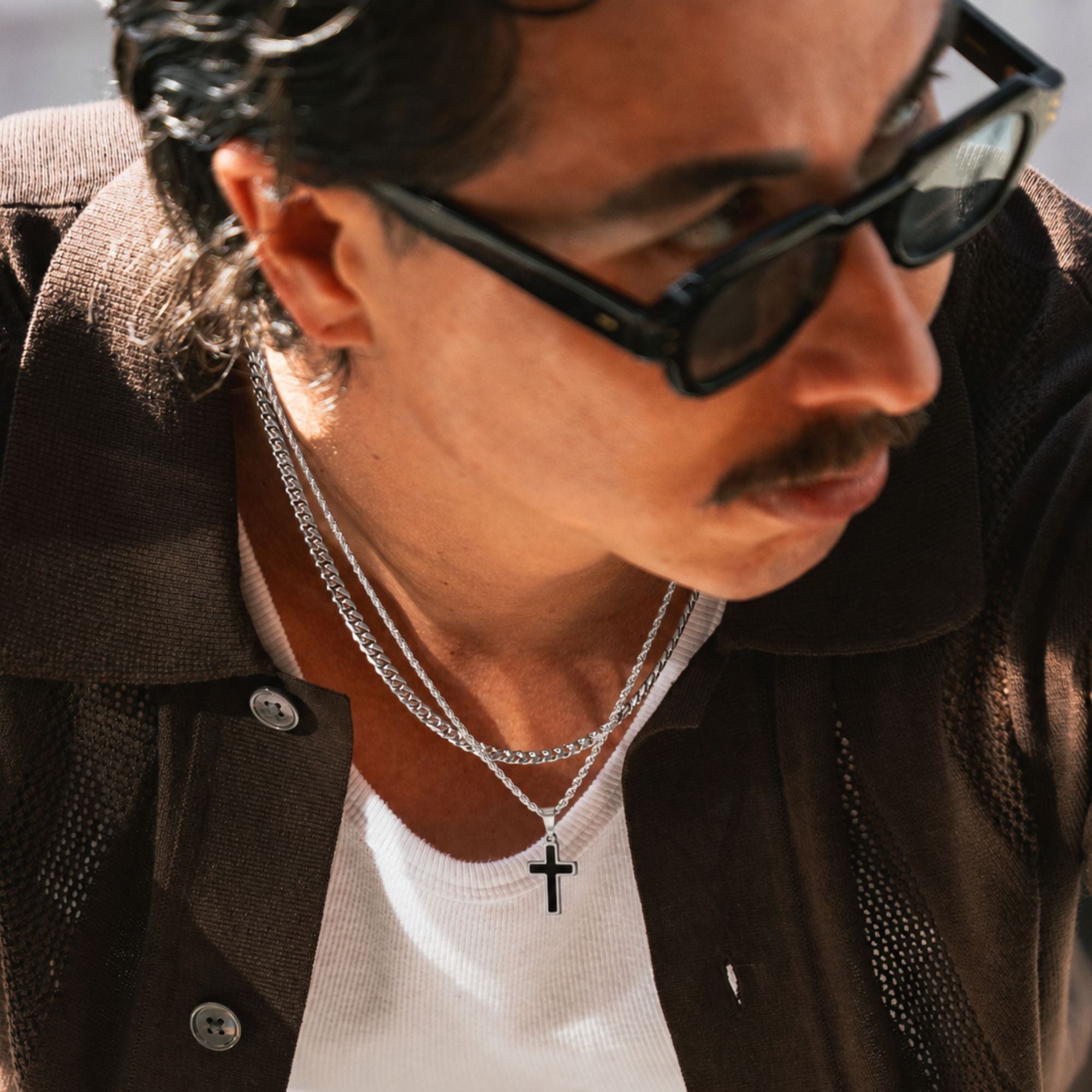 A close-up lifestyle shot of a man wearing a silver double-layer chain necklace with a small black cross pendant, styled with black sunglasses, a white t-shirt, and a brown jacket.