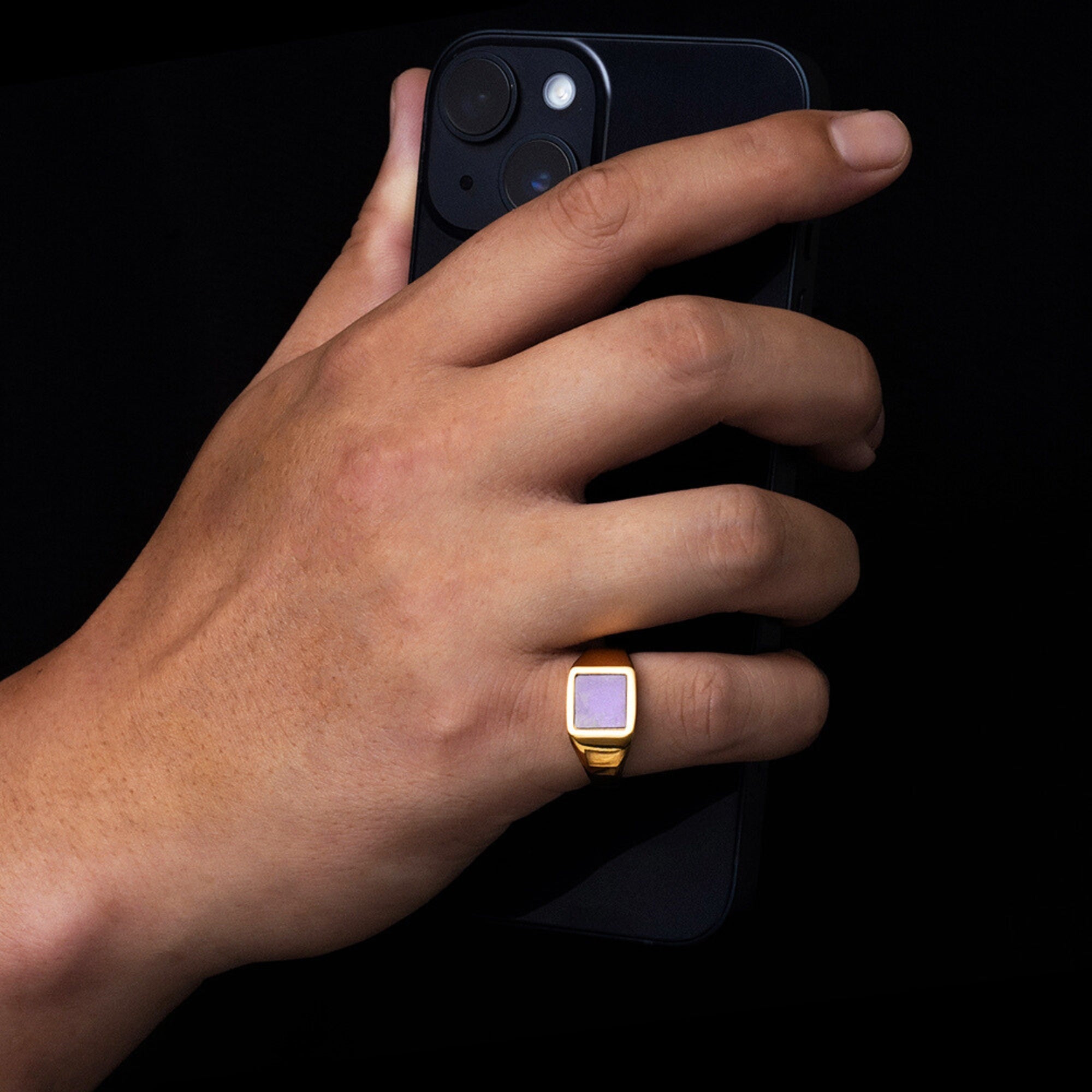 Hand holding a smartphone with a gold ring featuring a purple square stone on a black background