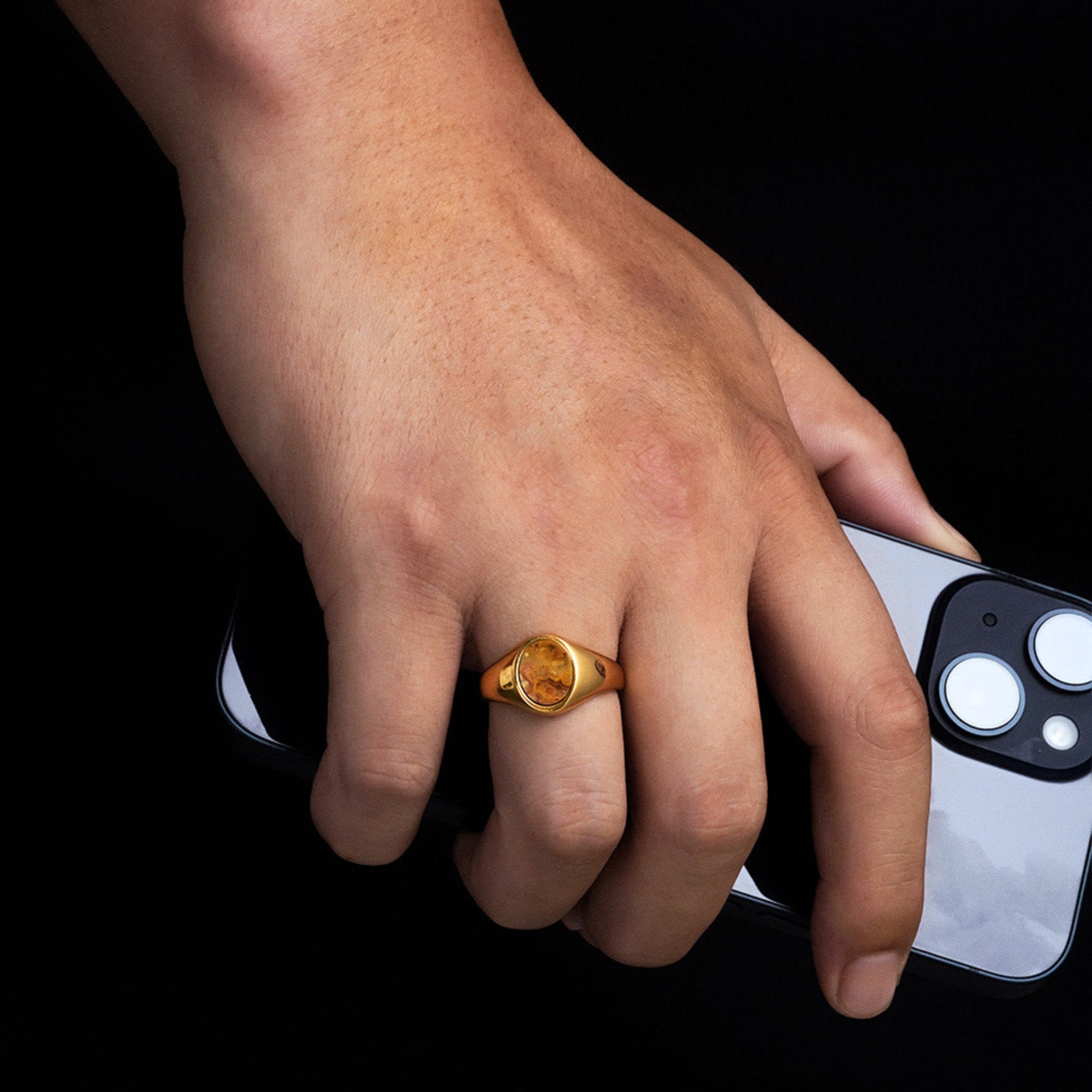Hand wearing a gold ring holding a smartphone with a black background