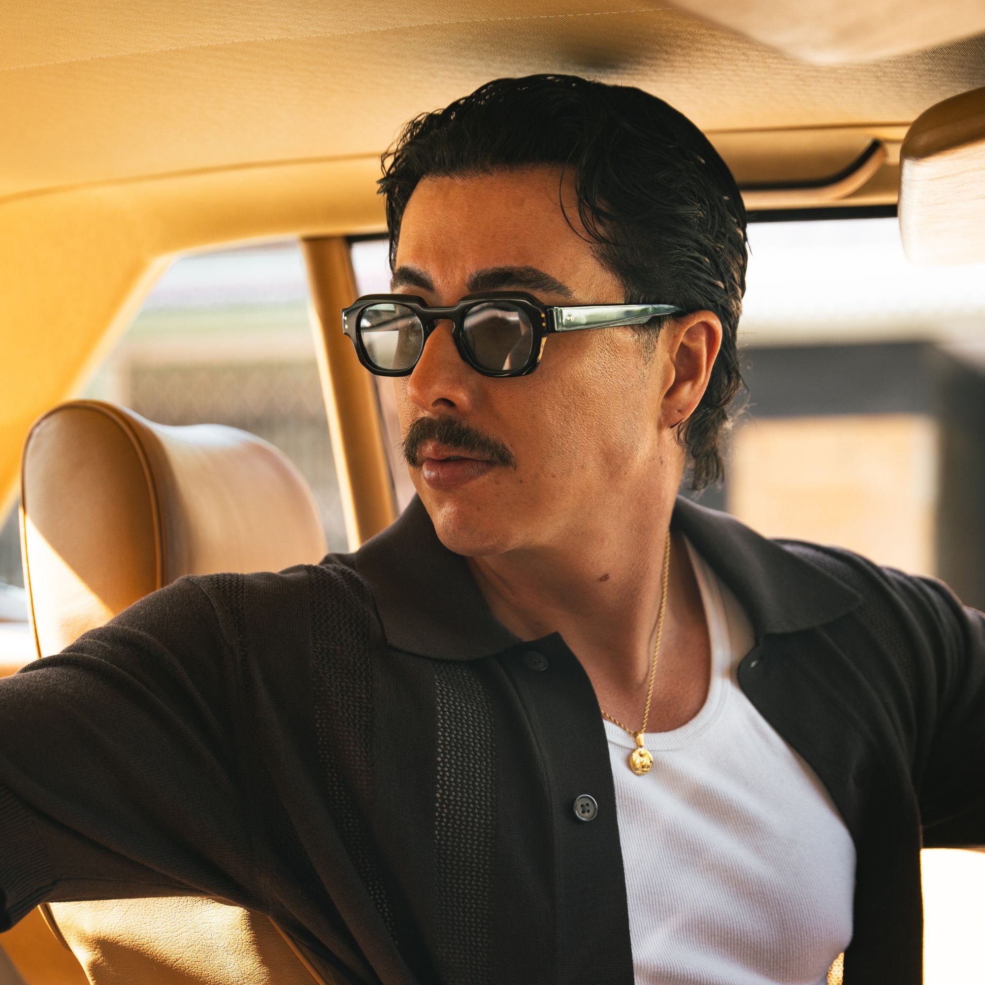 A man with a mustache wearing black sunglasses with green temples, a black knit polo shirt over a white t-shirt, and a gold necklace, sitting inside a car with tan leather seats.