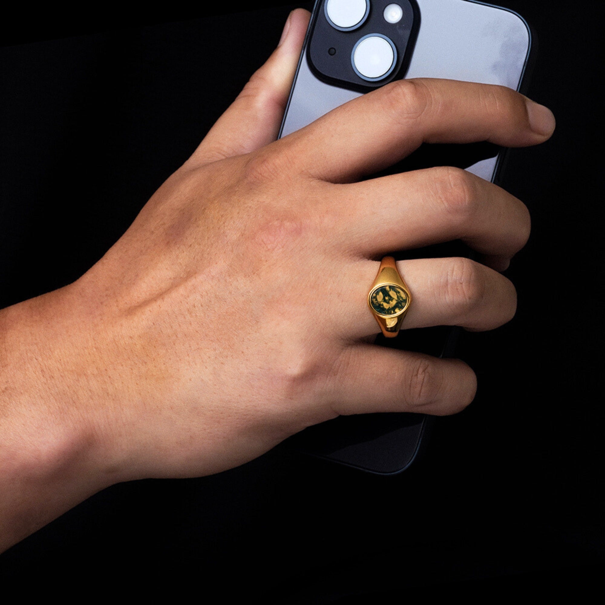 Hand wearing a gold ring holding a smartphone on a black background