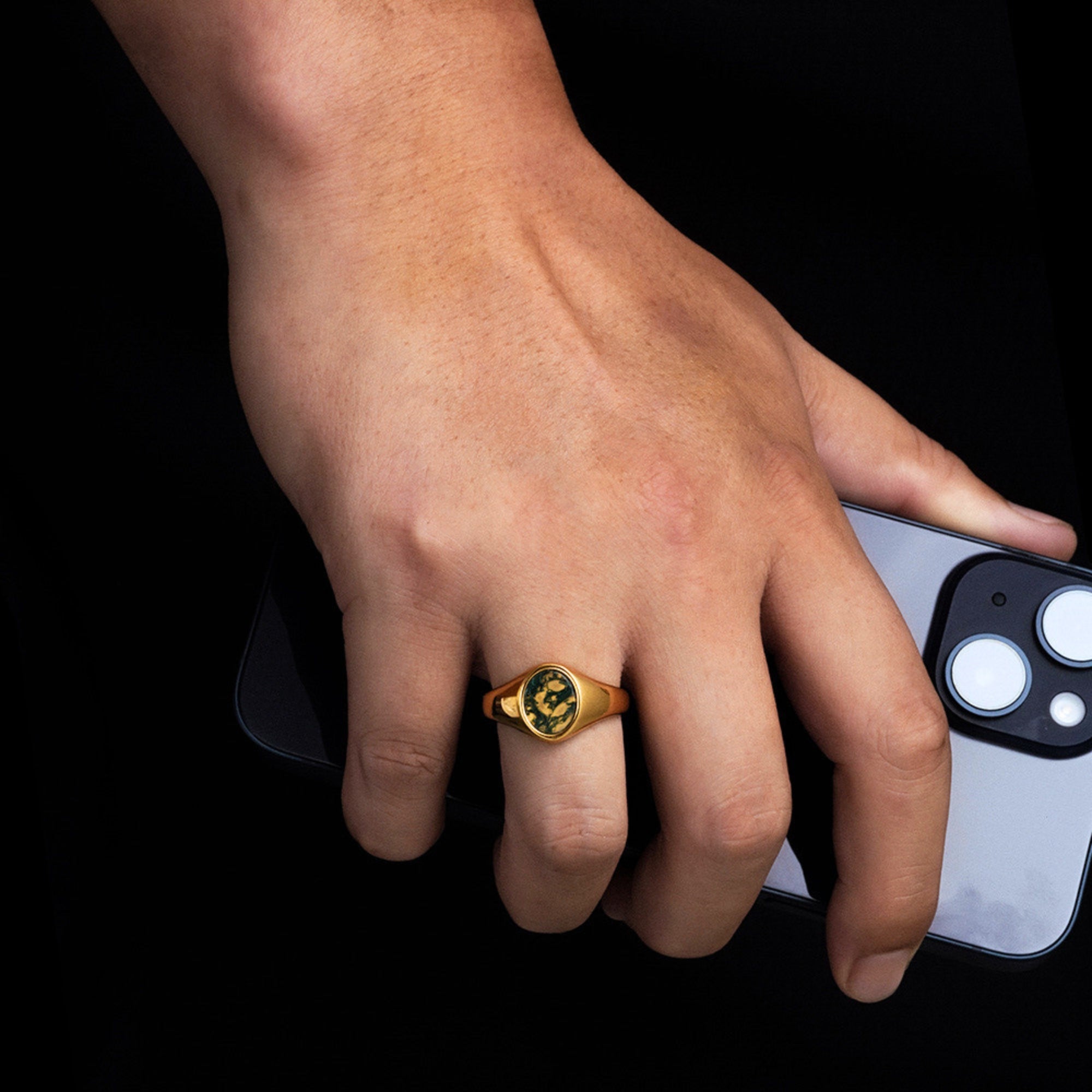 Hand wearing a gold ring holding a smartphone on a black background