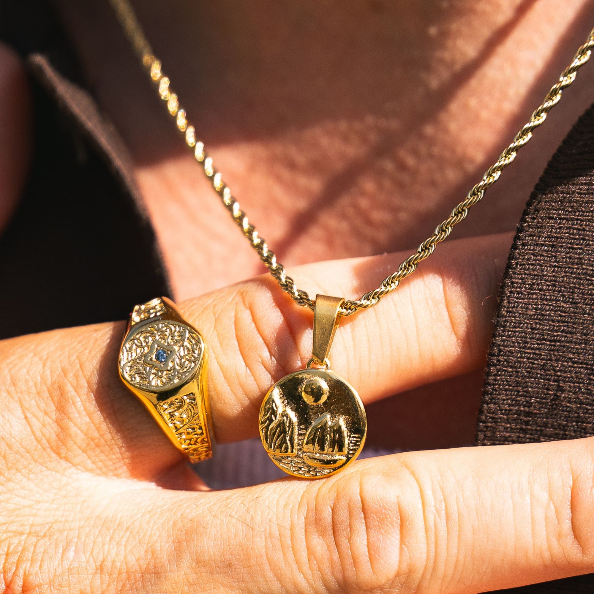 A close-up lifestyle shot of a hand holding two gold jewelry pieces: a round engraved gold pendant on a chain and a gold signet ring with an oval face and a small blue stone, set against a background of skin and a dark shirt.