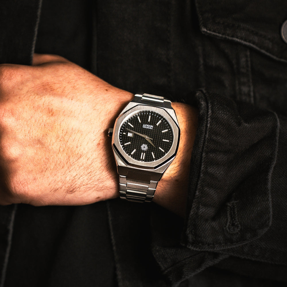 Person wearing a luxury watch with a black face and silver case on a dark background