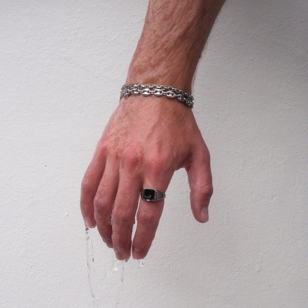 Hand wearing a silver chain bracelet and black ring on a plain background