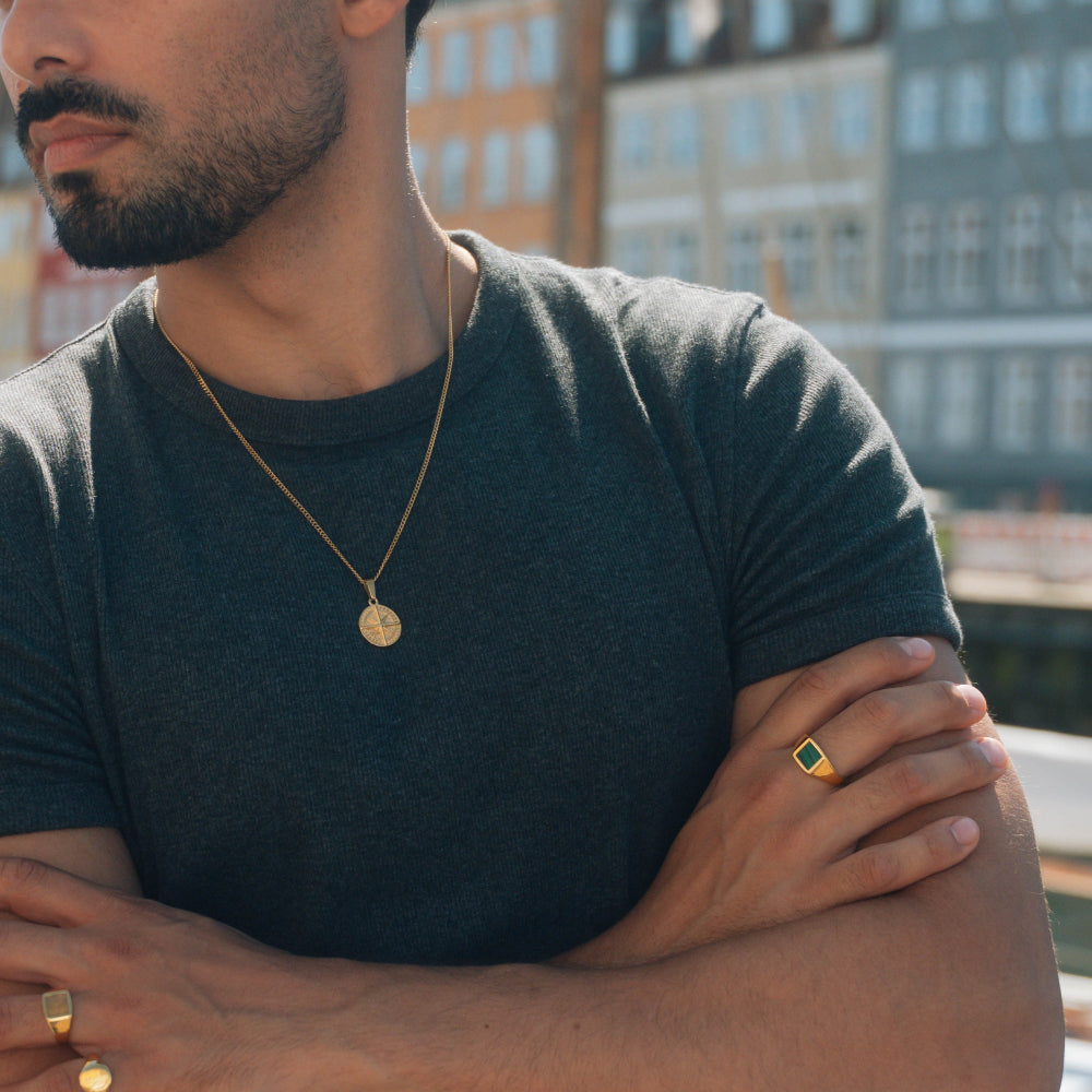 Man wearing a dark green t-shirt with gold jewelry, standing against a blurred cityscape.