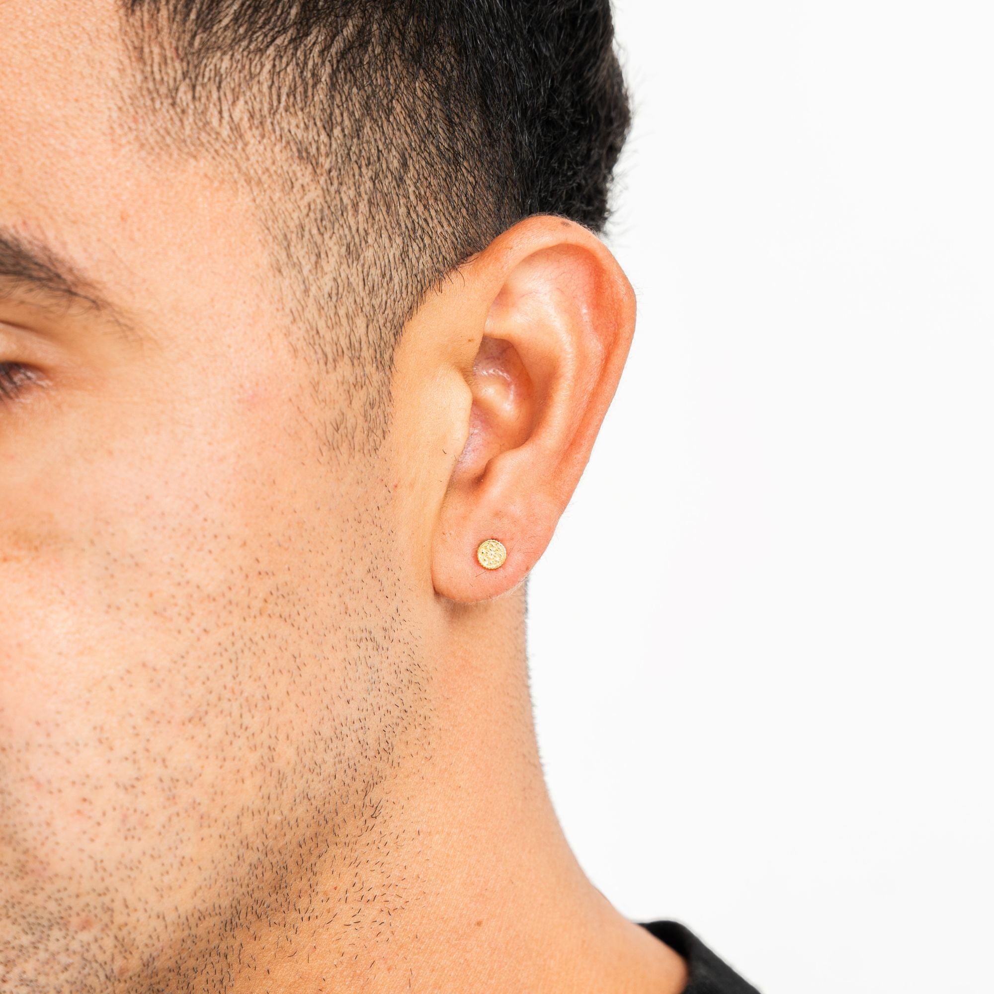 A close-up side profile of a man's head showing a small, round gold stud earring in his earlobe against a clean white background.