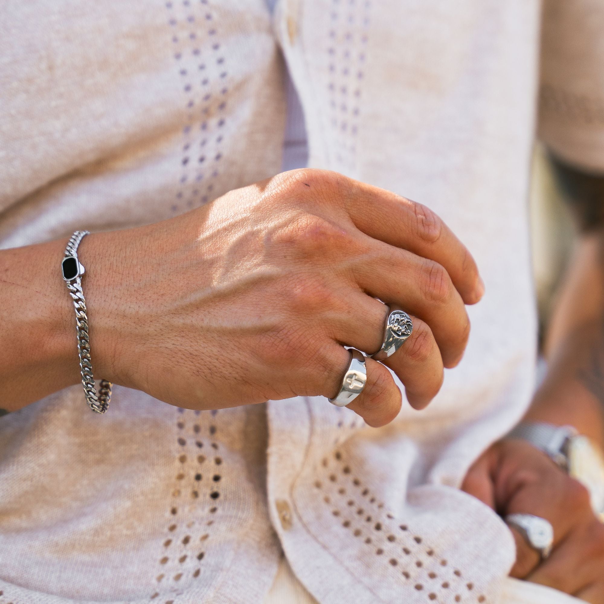 A close-up of a person's hand wearing a silver chain bracelet with a black square accent and silver rings, set against a light beige textured knit cardigan.