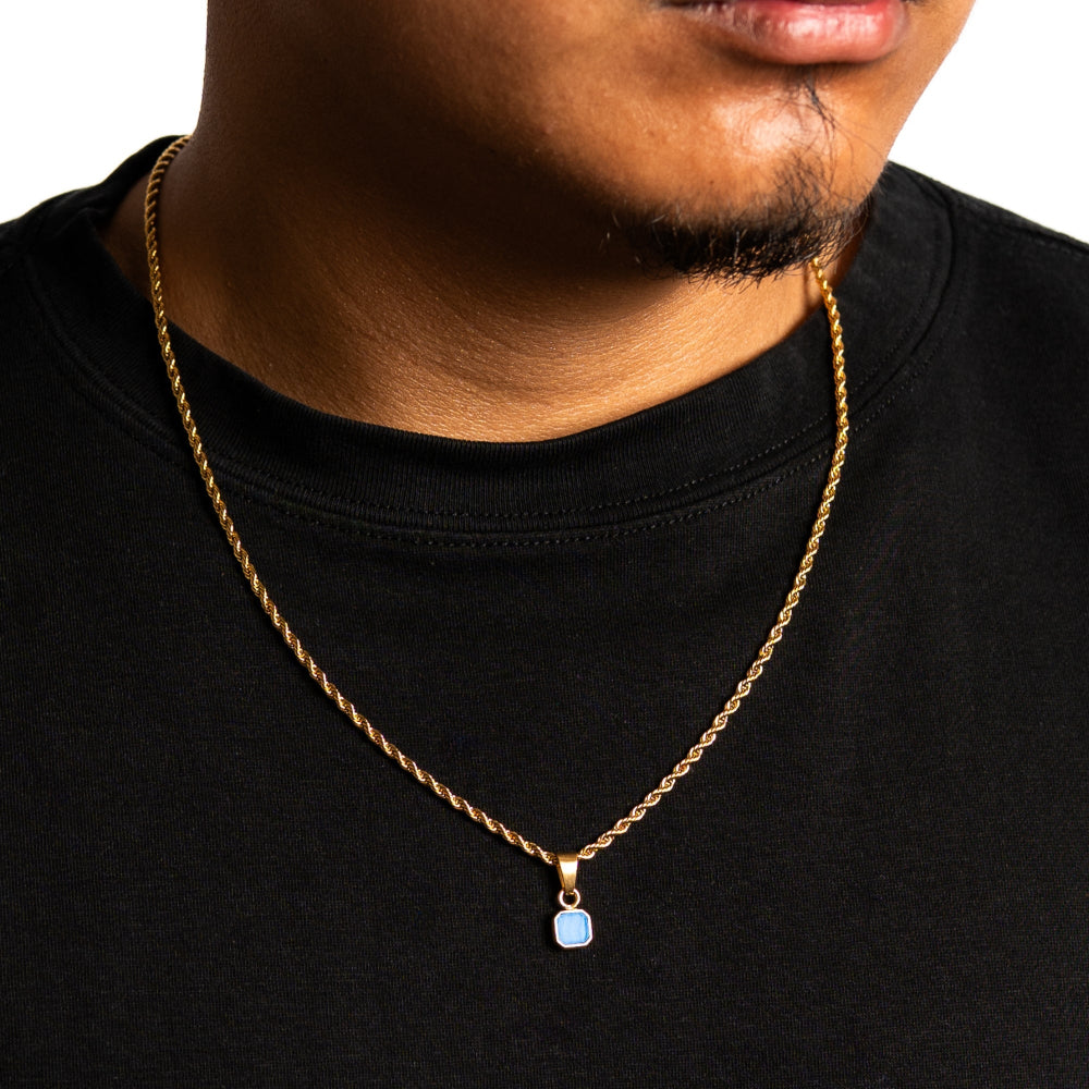 Man wearing a gold chain necklace with a blue pendant on a black shirt.