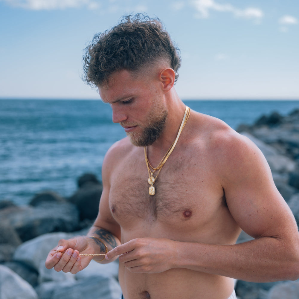 Man standing on a rocky beach with ocean view, wearing a gold necklace.