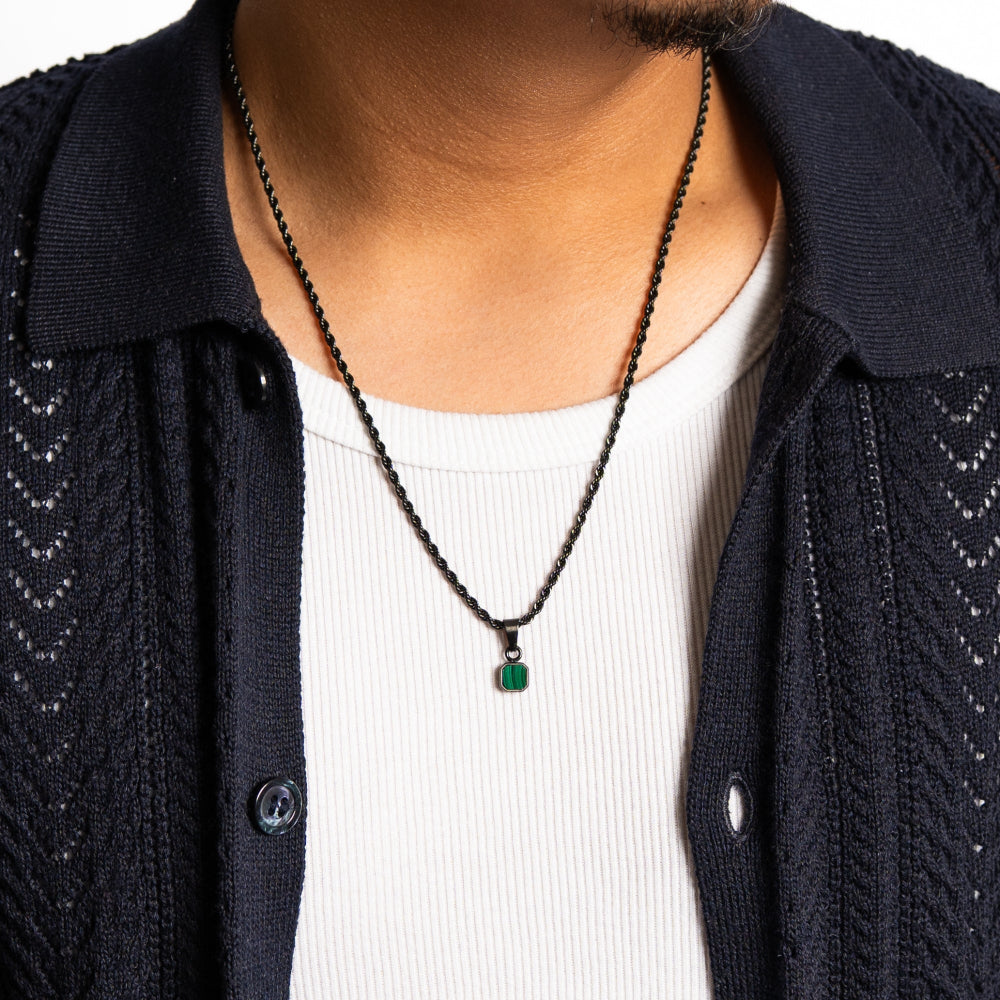 Person wearing a black chain necklace with a green pendant, dressed in a dark jacket and white shirt.