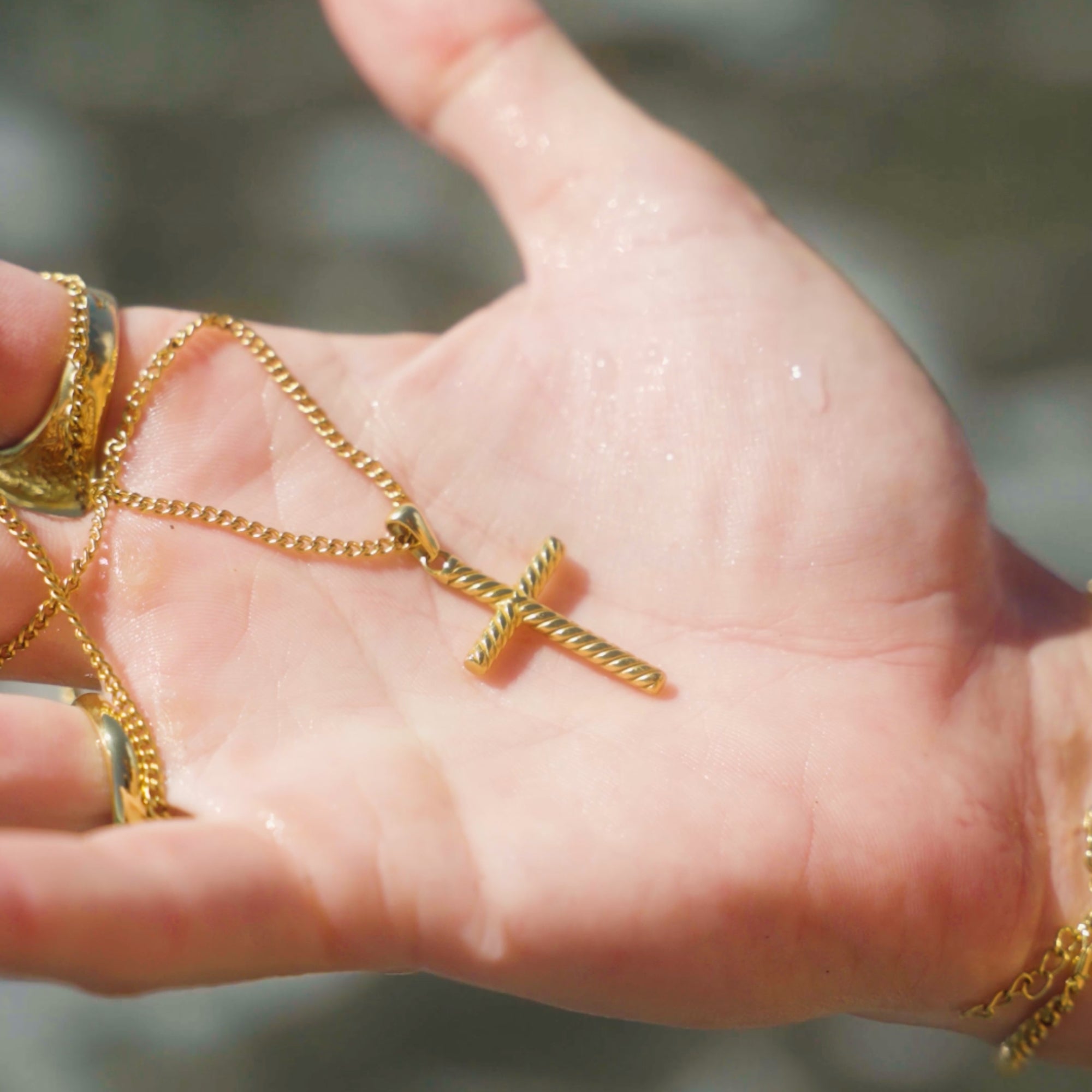 Hand holding a gold necklace with a cross pendant against a blurred natural background