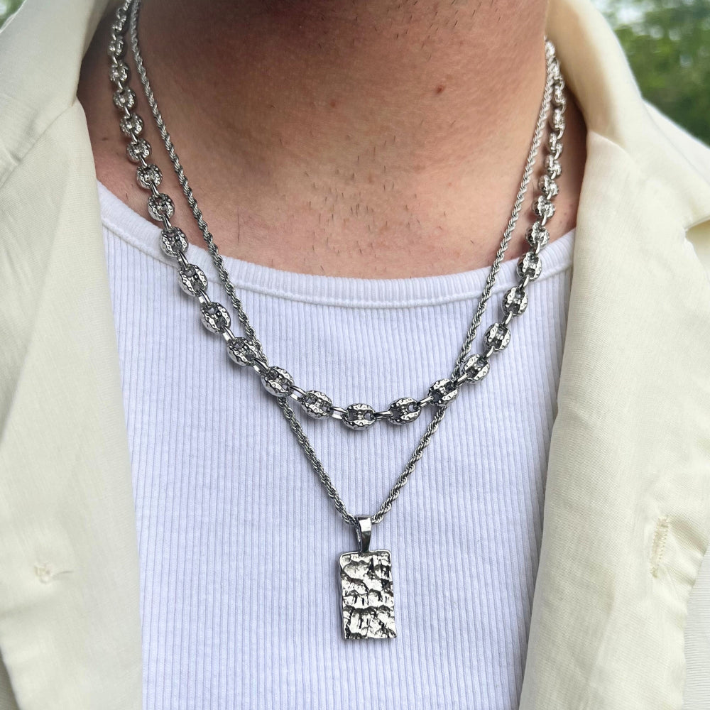 Person wearing a silver chain necklace with a textured pendant on a white shirt.