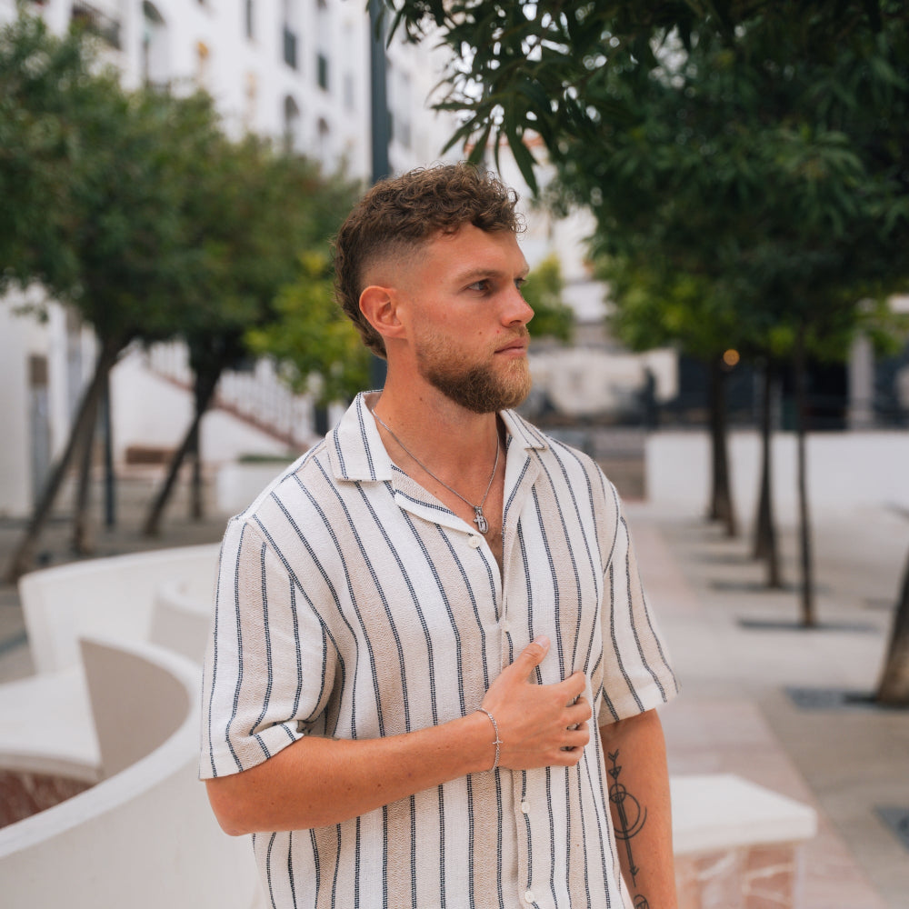 Man wearing a striped shirt standing outdoors with trees and buildings in the background