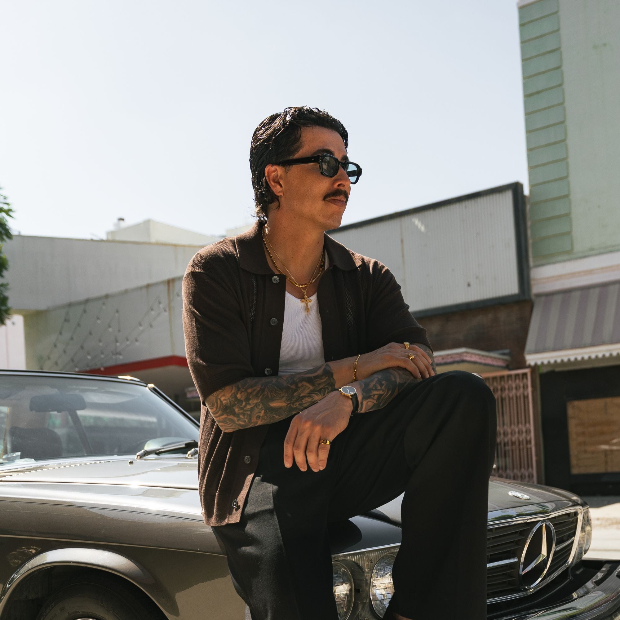 A man with a mustache and tattoos wearing a dark brown button-up shirt, black pants, and sunglasses, sitting on the hood of a classic Mercedes-Benz car in an outdoor urban setting.