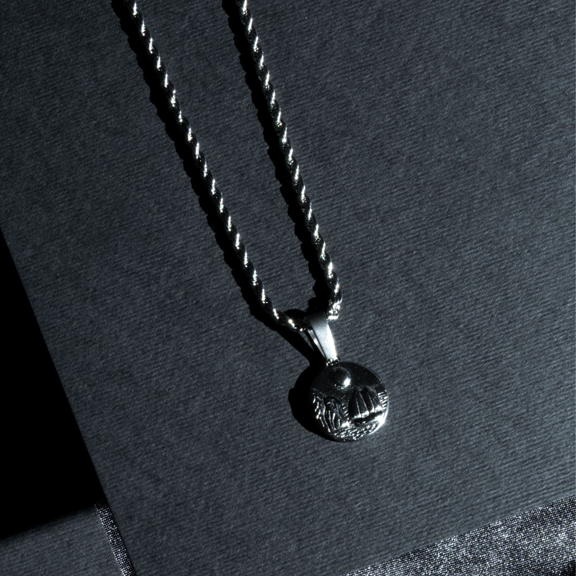 A silver-toned twisted rope chain necklace with a textured circular pendant, displayed on a dark gray textured background.