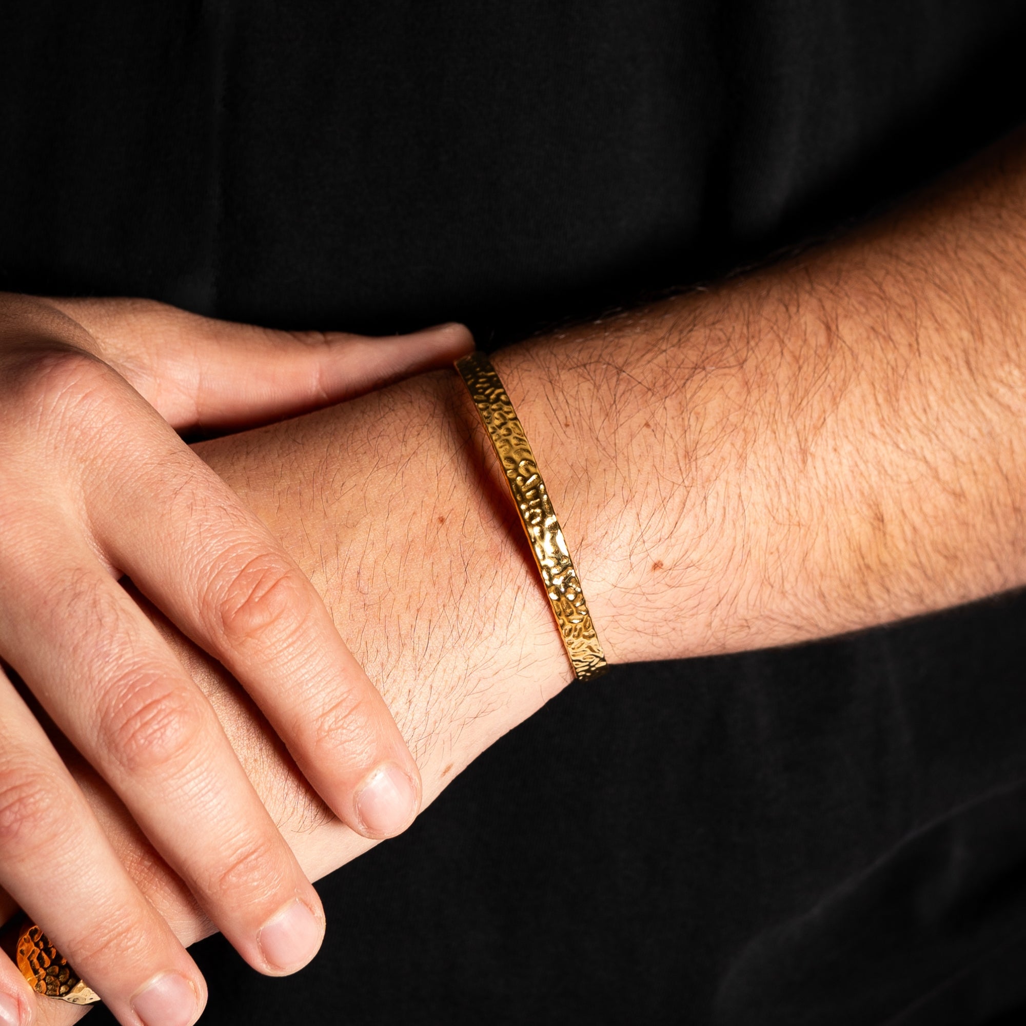 Gold bracelet on a wrist with a hand resting on it, against a black background