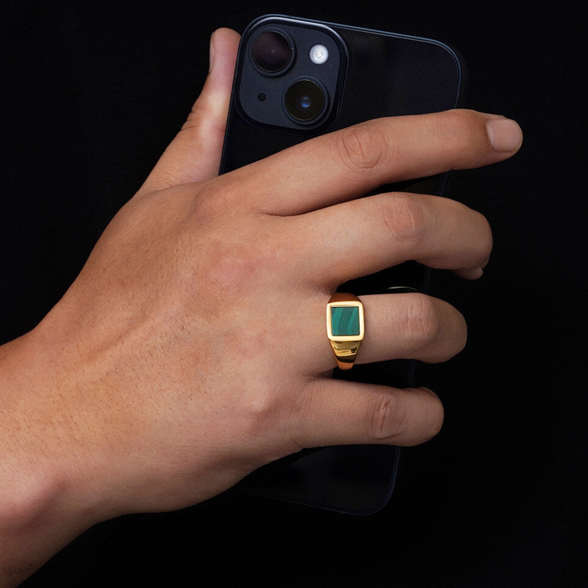 Hand holding a black phone with a gold ring featuring a green gemstone on a black background