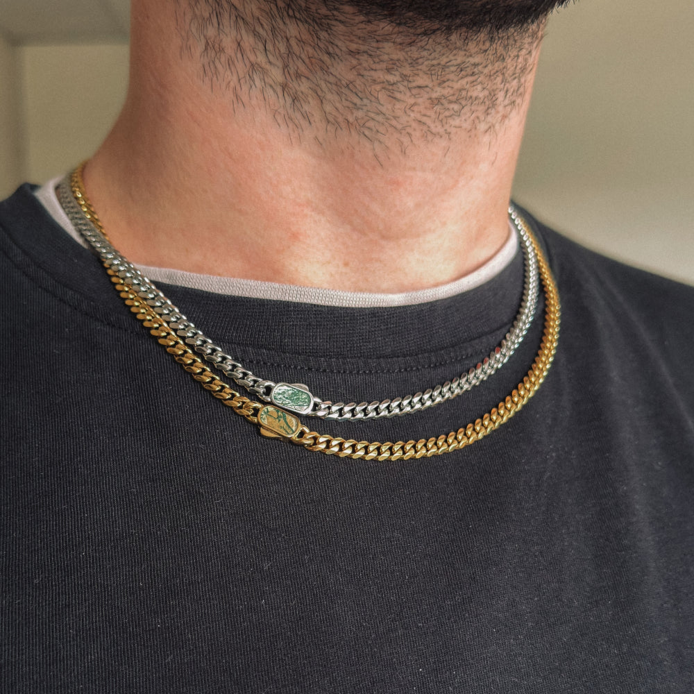 Close-up of a person wearing two gold chain necklaces with green gemstones on a plain background.