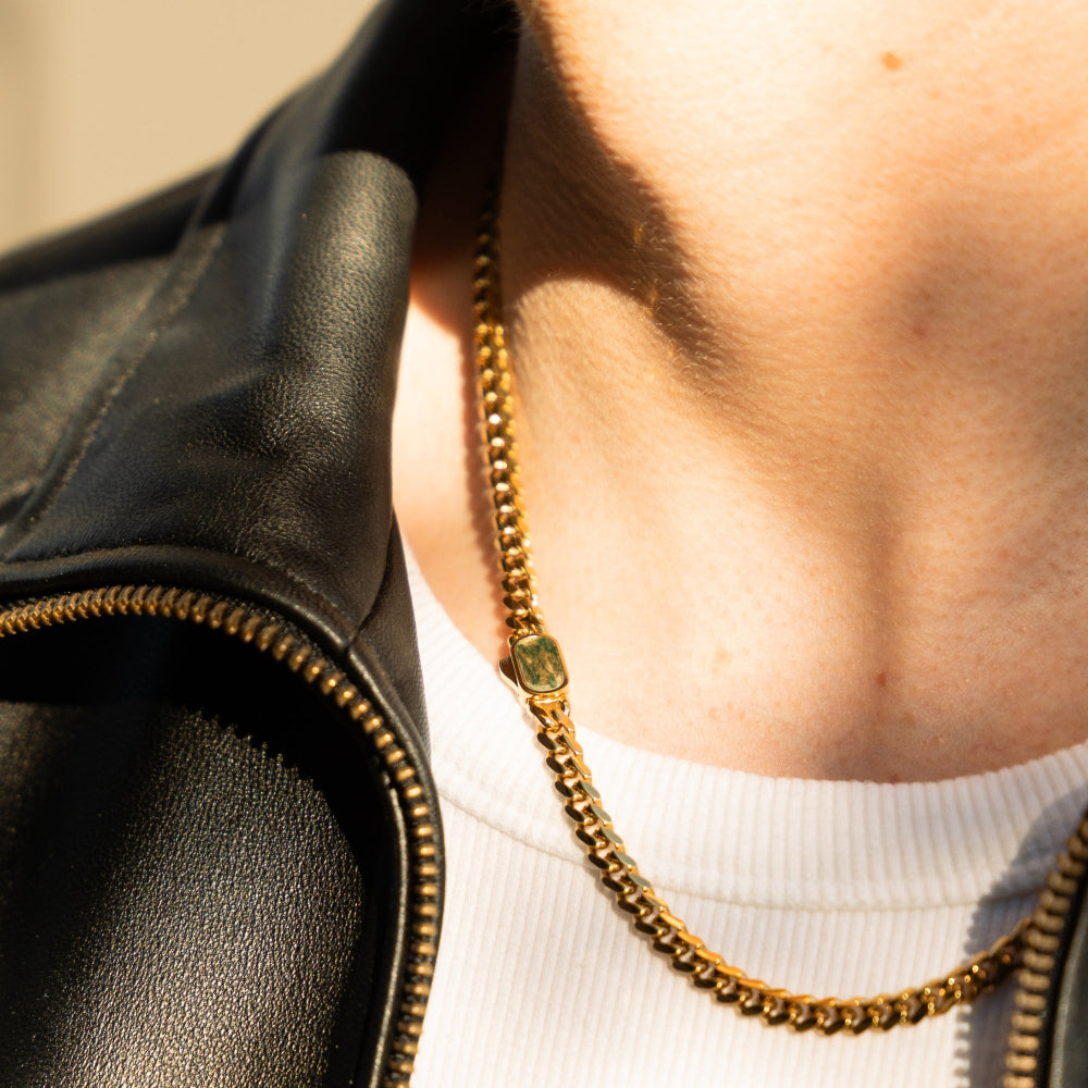 Person wearing a black leather jacket and gold chain necklace with a green pendant.