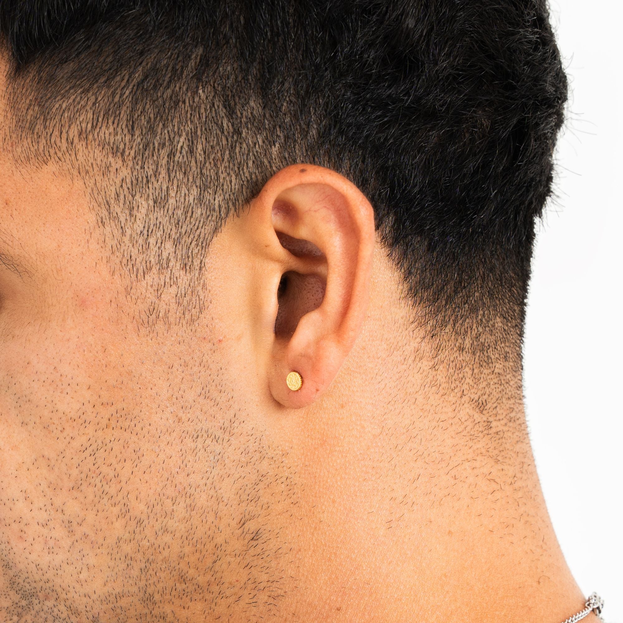 A close-up of a man's ear wearing a small, round gold stud earring against a clean white background.