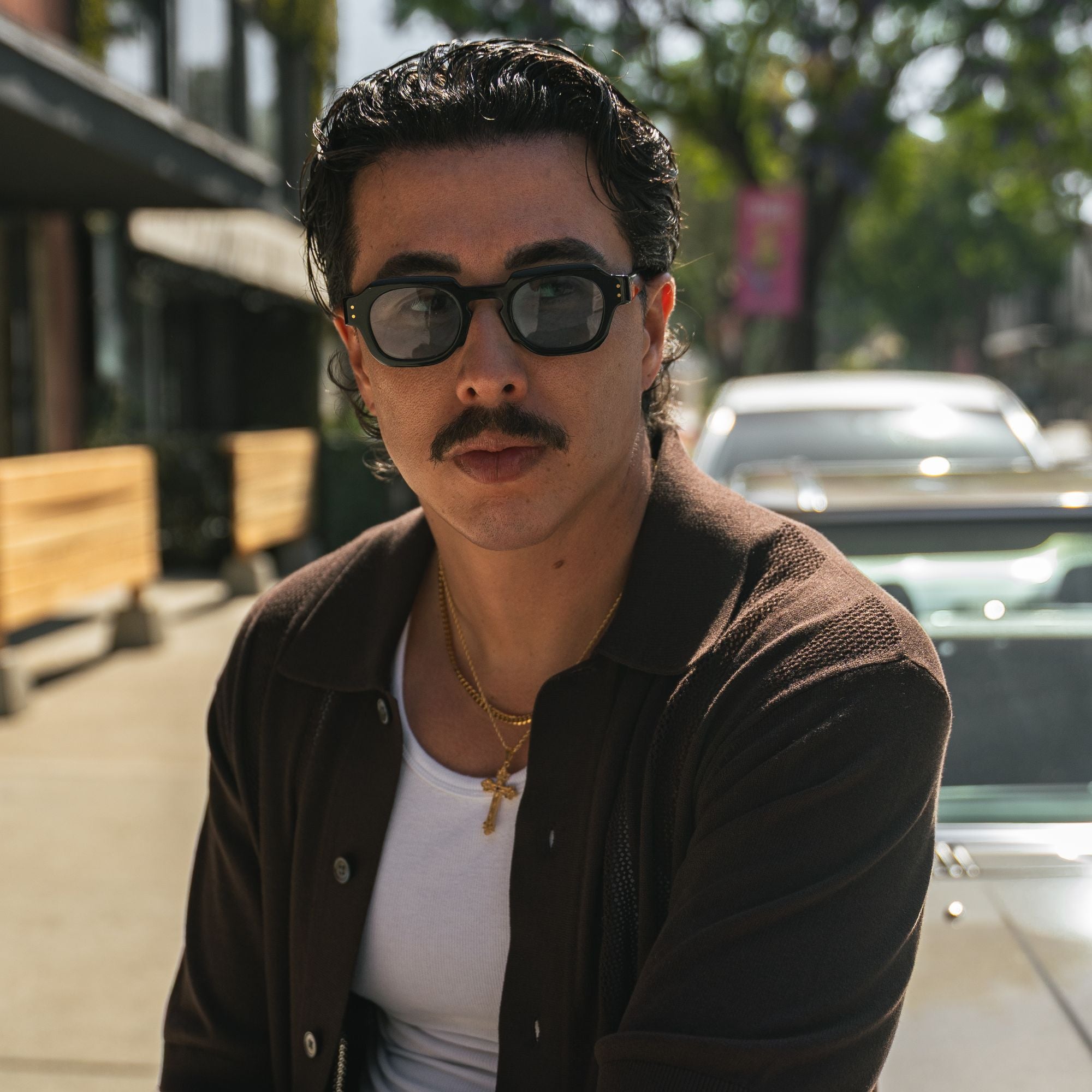 A man with a mustache wearing black sunglasses, a brown jacket, and a white tank top, photographed outdoors on a sunny street with cars and trees in the background.