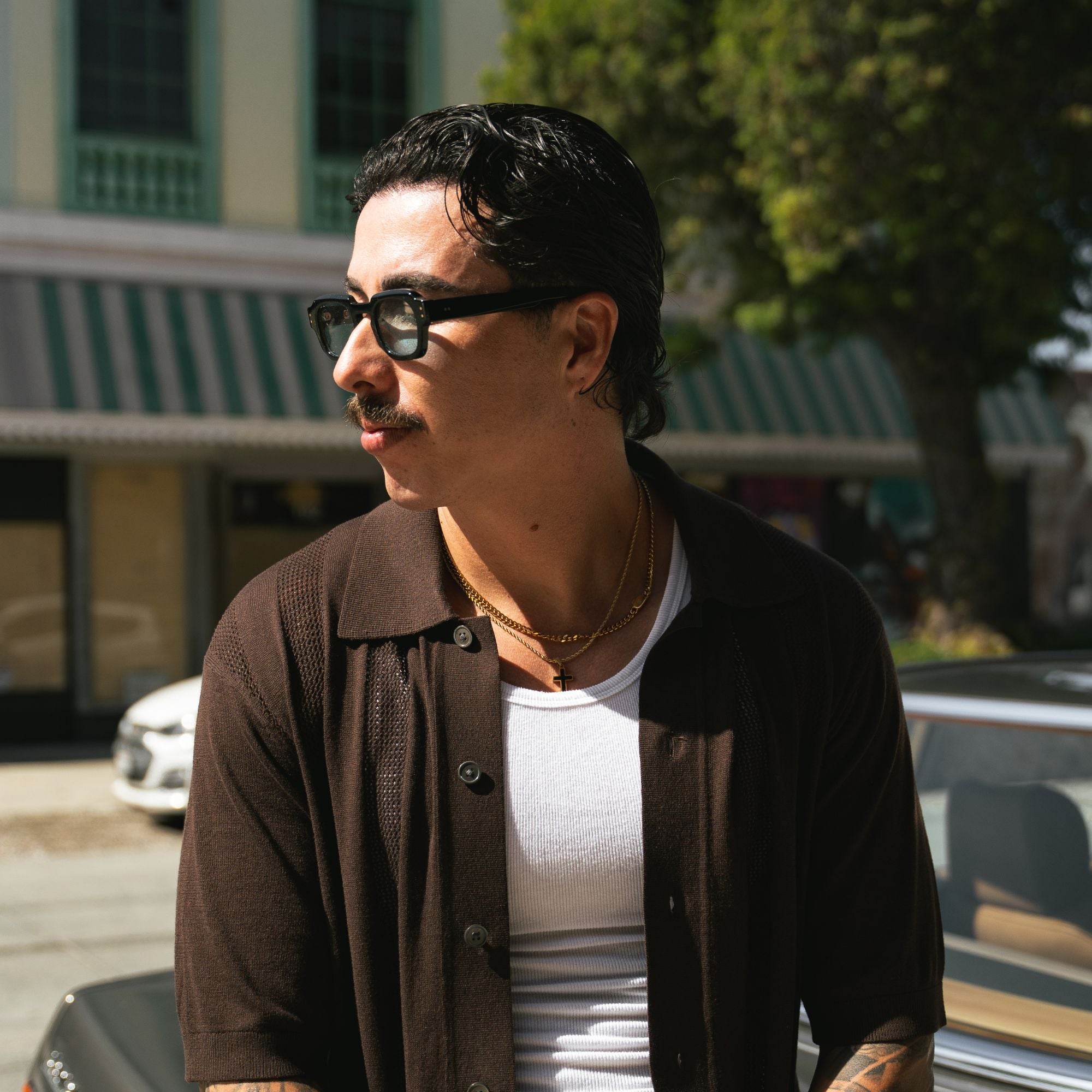 A man wearing black sunglasses, a brown textured button-up shirt over a white t-shirt, and gold necklaces, standing outdoors in front of a building with green awnings and trees.