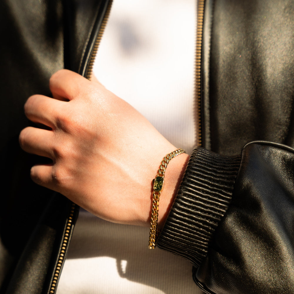Hand wearing a gold bracelet with a green gemstone, holding a black leather jacket.