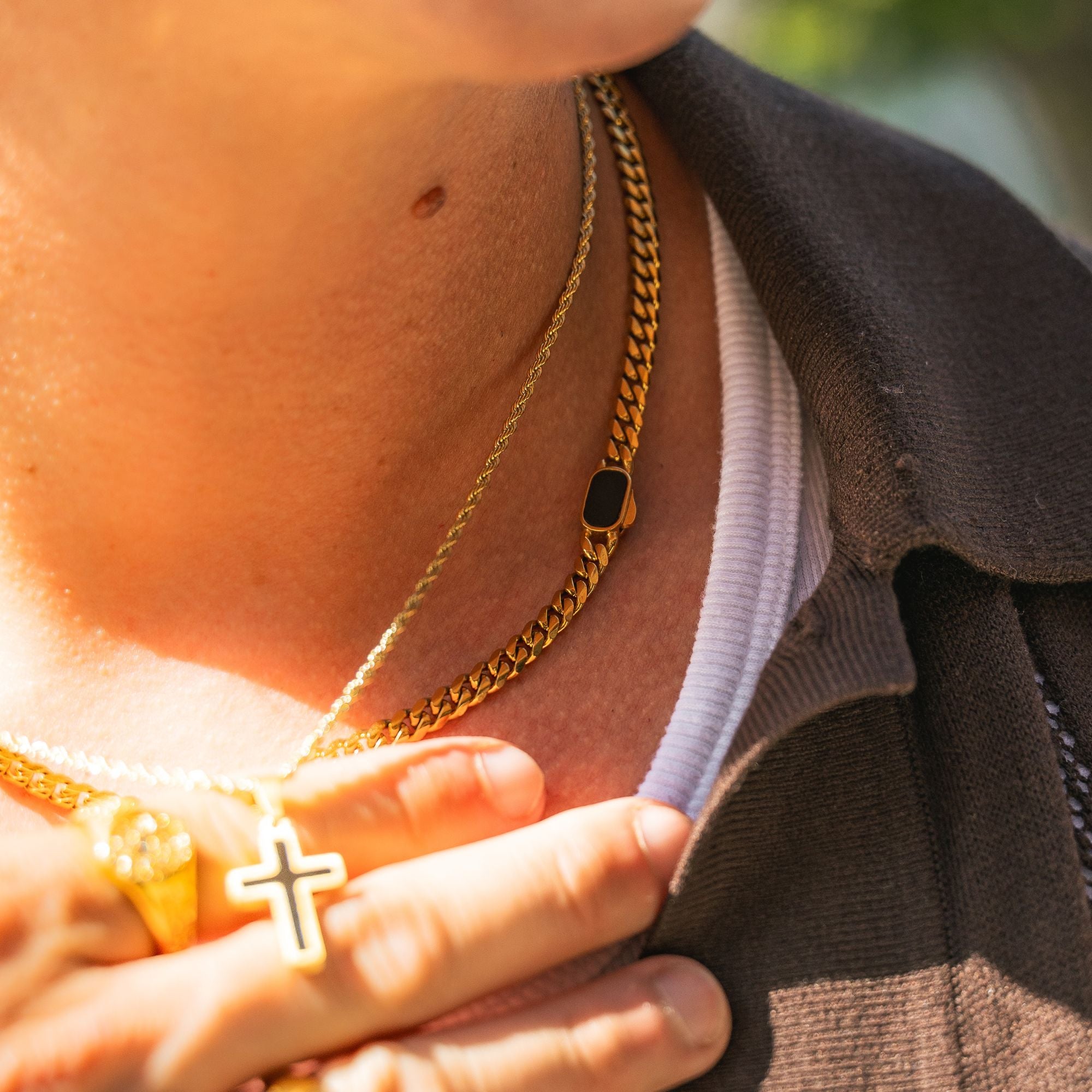 Close-up of a person wearing layered gold necklaces, including a curb chain with a small black rectangular accent and a cross pendant, over a white shirt and dark brown jacket.