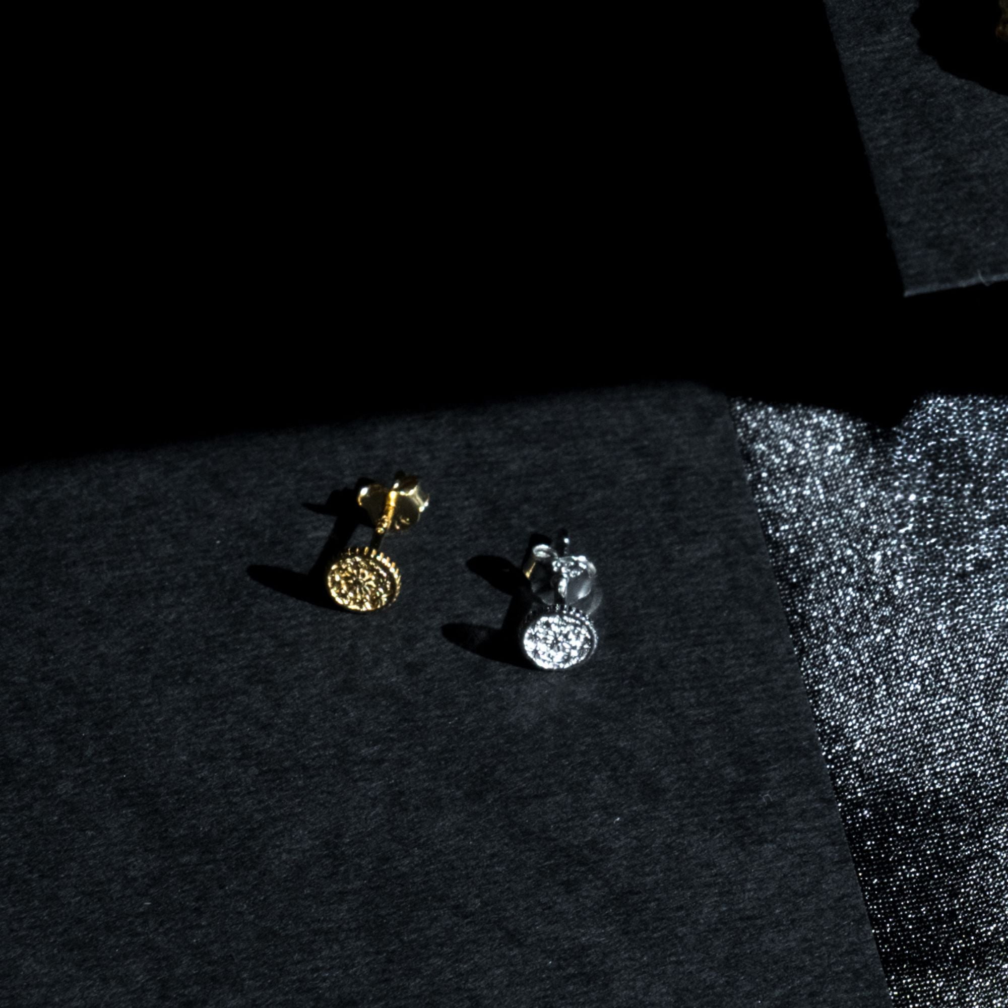 Two small stud earrings on a dark textured surface, one gold-toned with a circular textured face and one silver-toned with a faceted clear stone face, photographed in dramatic low light.