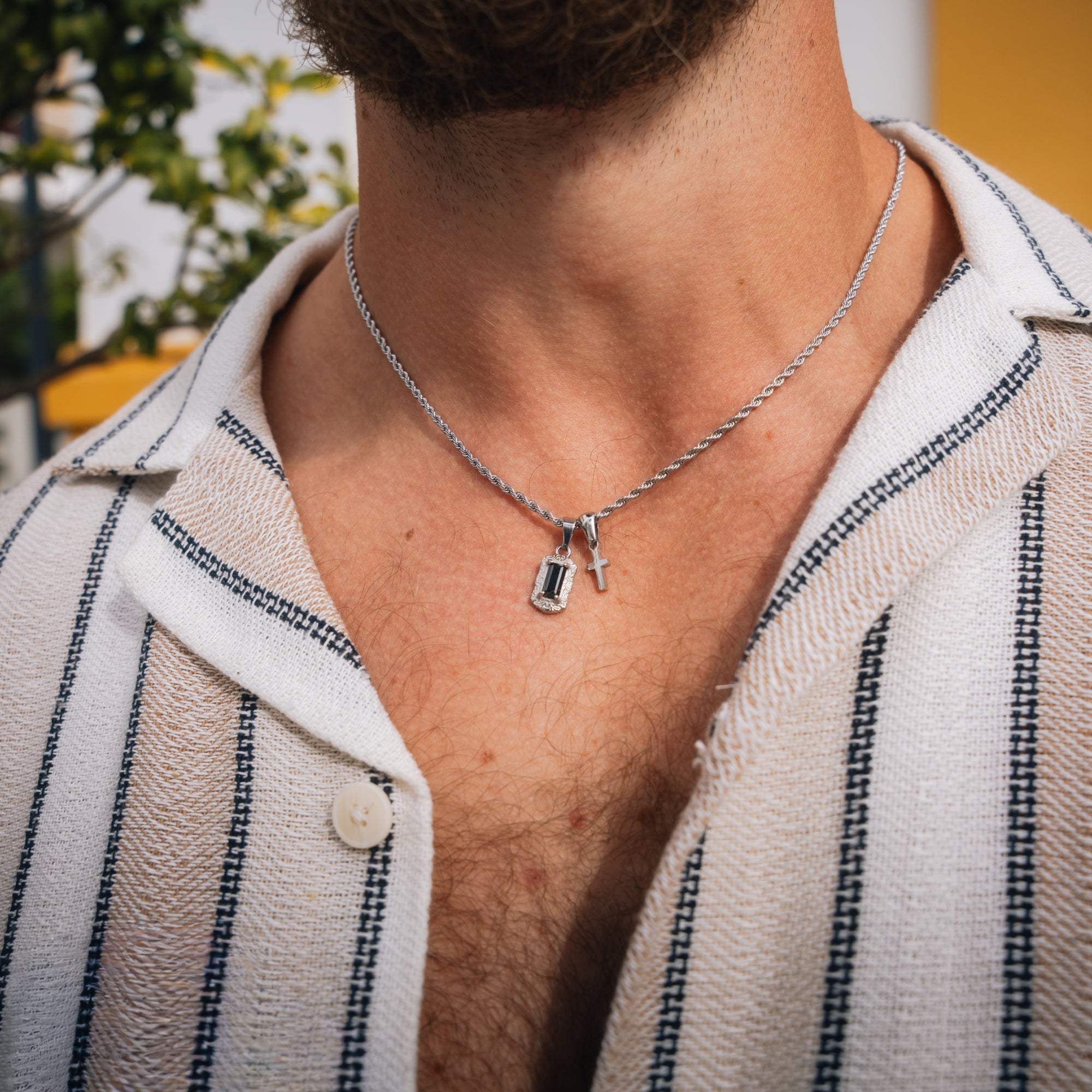 Man wearing a silver necklace with a pendant, outdoors.