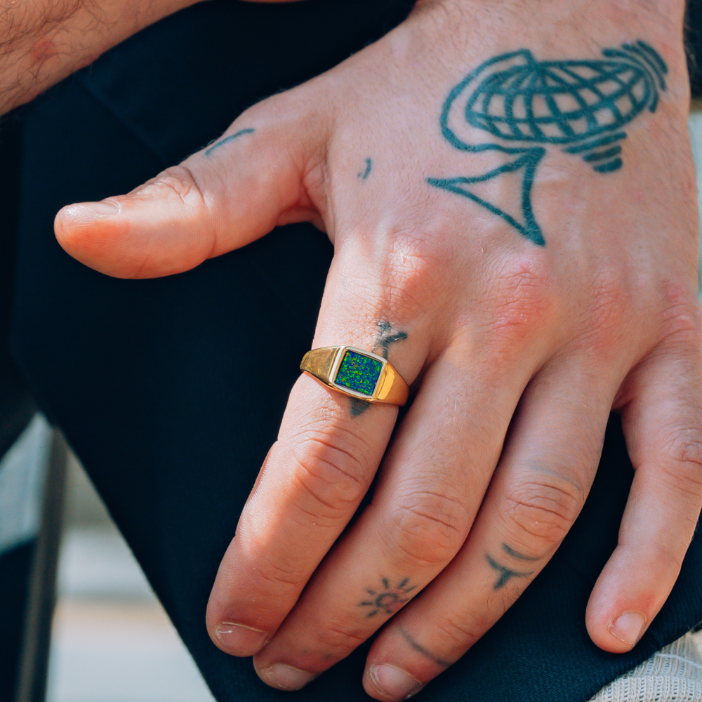 Hand with a gold ring featuring a green stone, surrounded by tattoos on a dark background